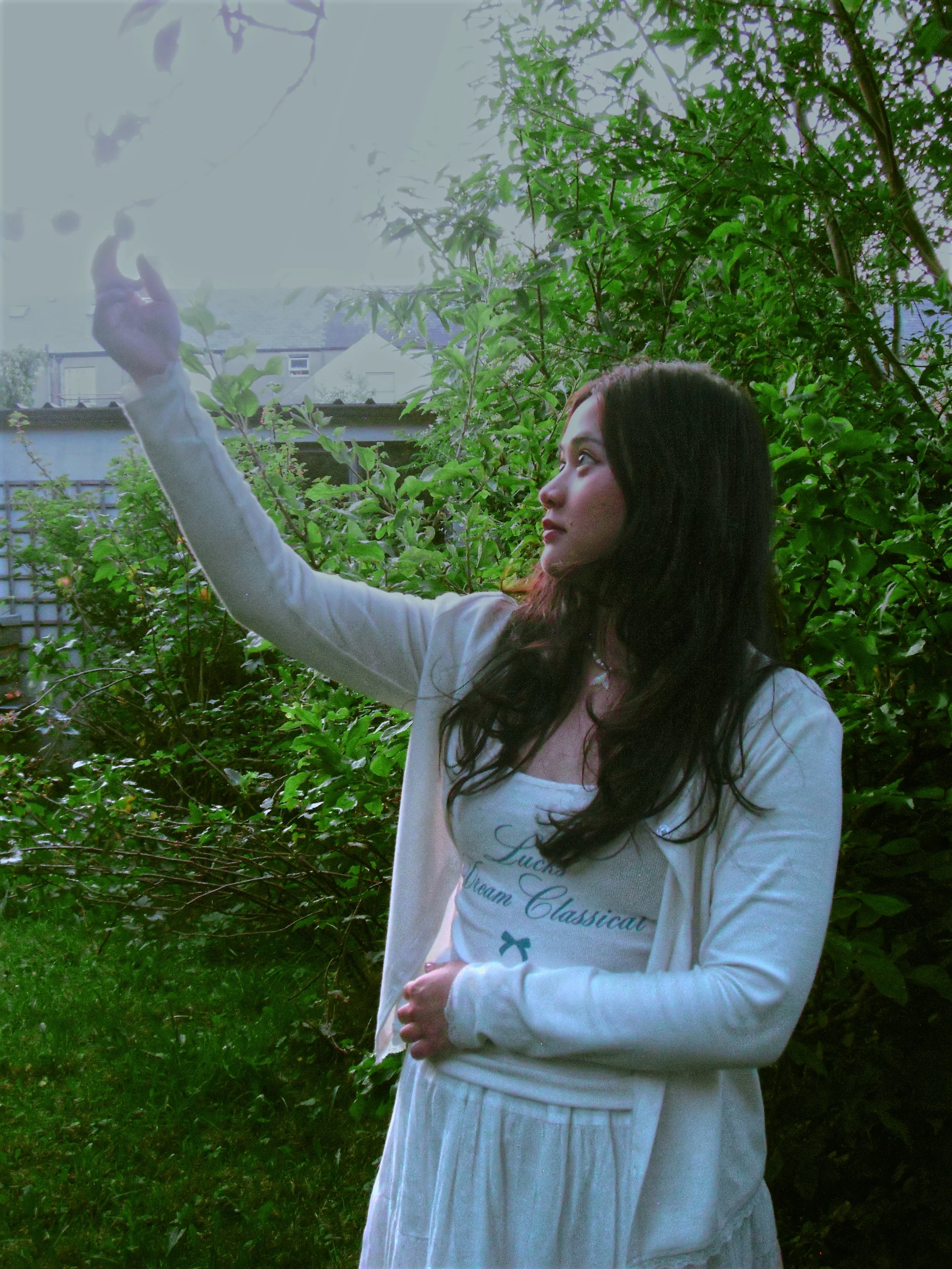 A young woman with long dark hair standing outdoors amidst green foliage, raising her right hand. She is wearing a light-colored cardigan over a white shirt and looking to her left.