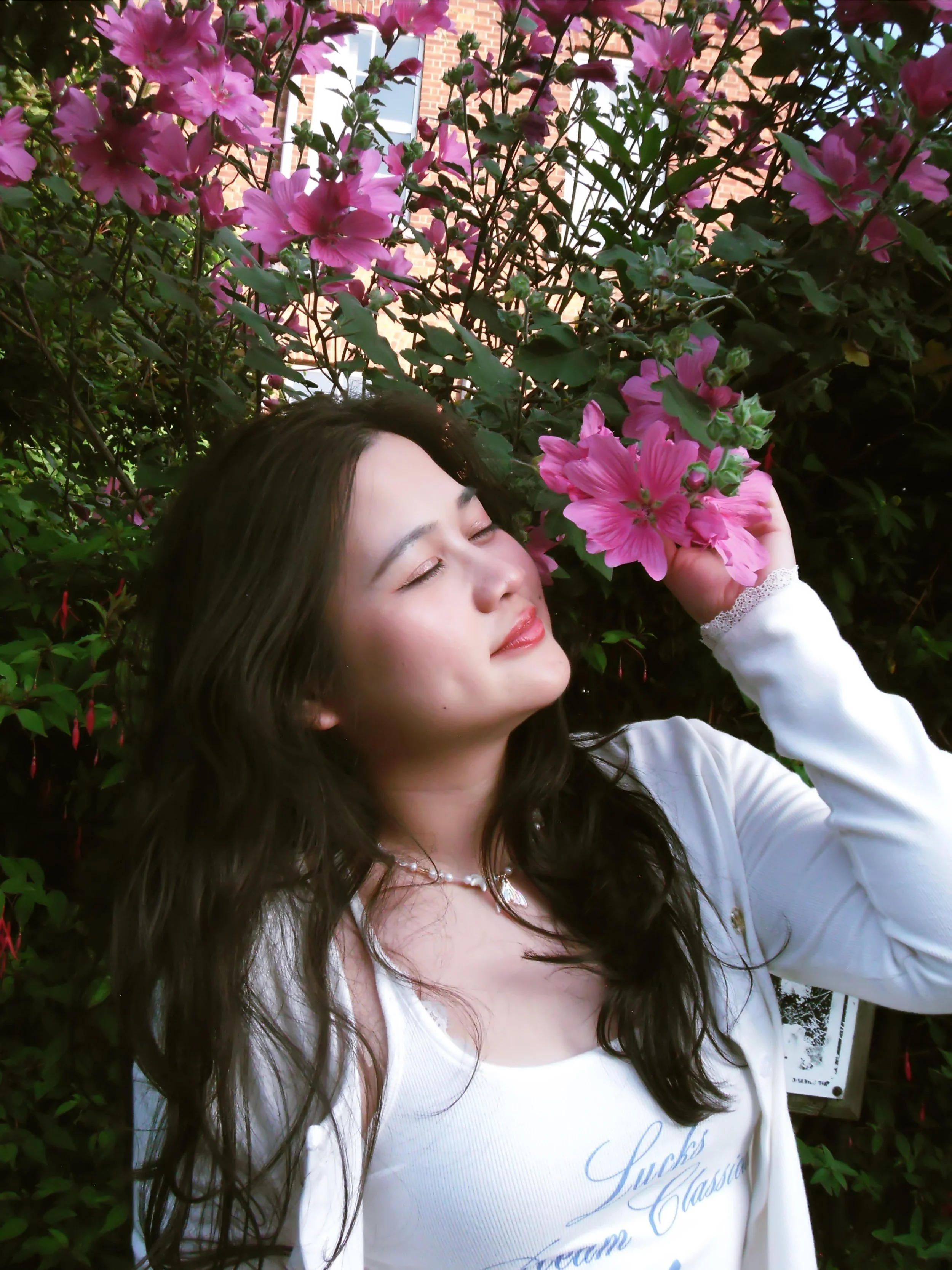 A young woman with long dark hair and light skin is standing outdoors among pink flowering bushes, gently holding some pink flowers near her face with her eyes closed and a peaceful expression.