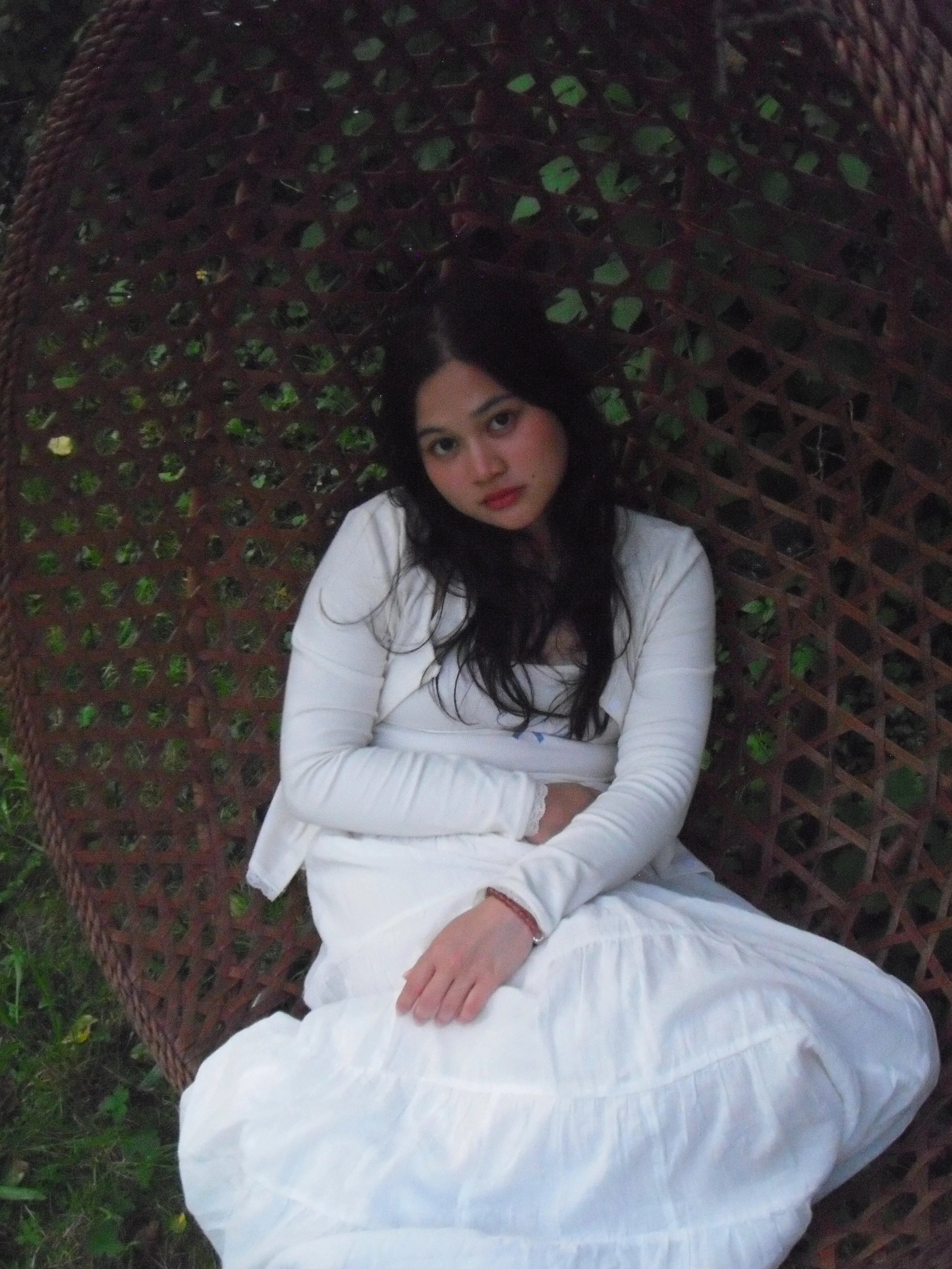 A woman with long dark hair, wearing a white dress and jacket, sitting on a round, wicker chair outdoors with greenery in the background.