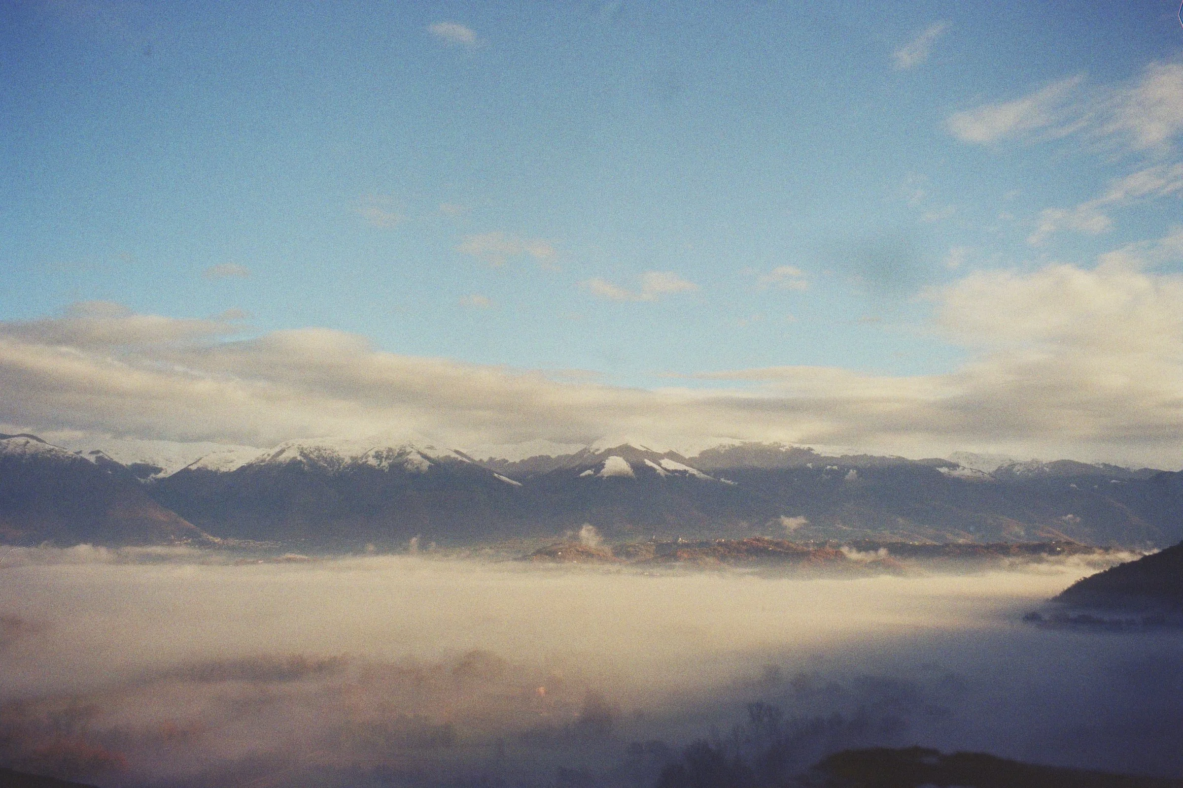 Mountain range with snow-capped peaks under a partly cloudy sky, overlooking a landscape shrouded in fog.