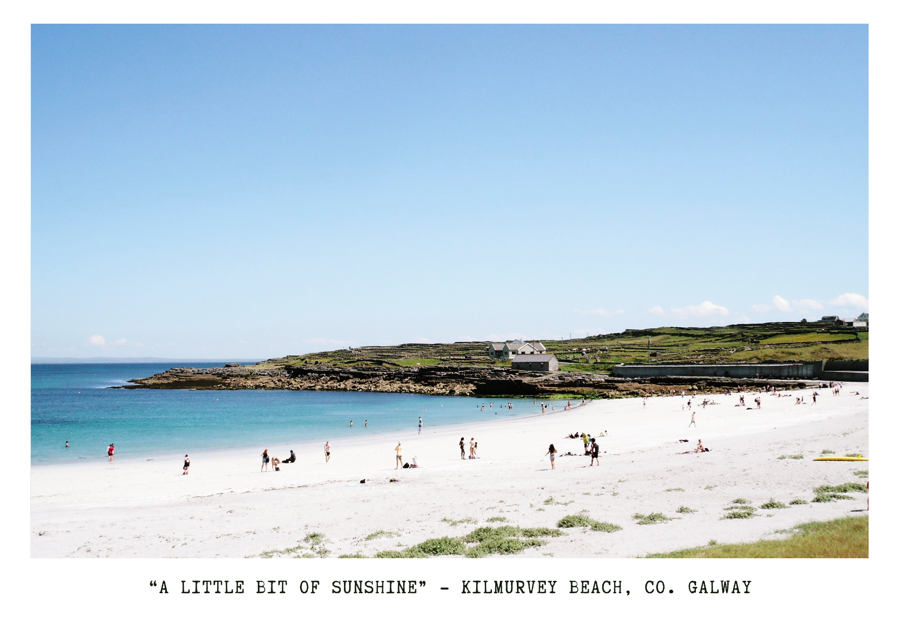 A scenic beach with white sand, colorful people, and calm blue waters, with a grassy hillside and white buildings in the background, under a clear blue sky.
