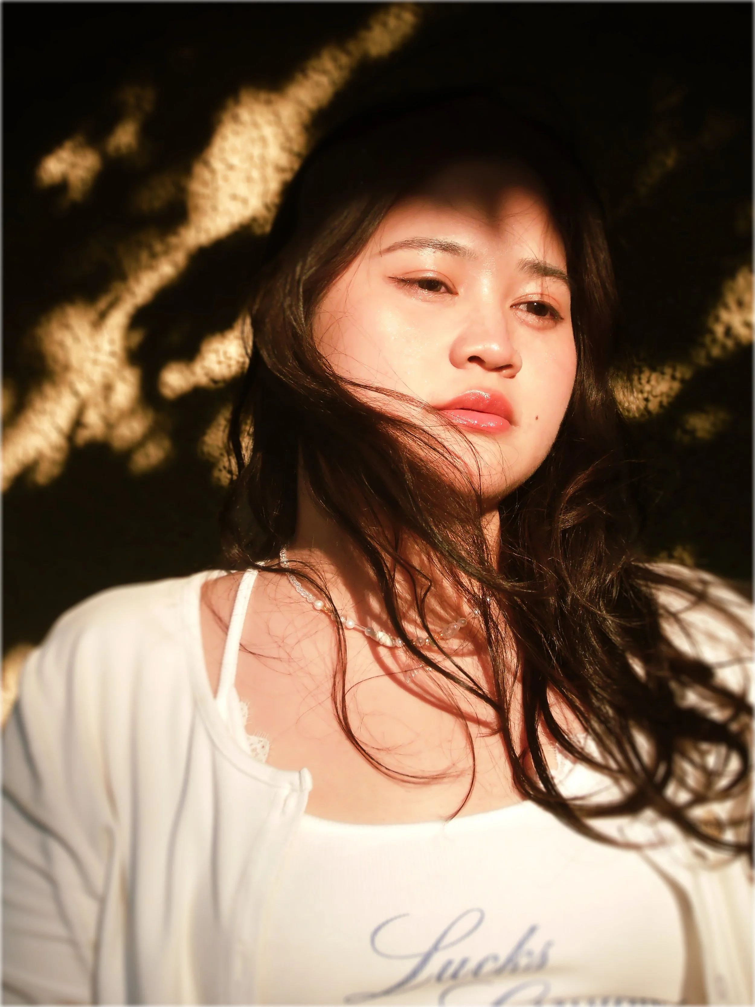 Close-up photo of a young woman lying on a black and beige patterned surface, wearing a white top and a pearl necklace, with soft lighting highlighting her face.