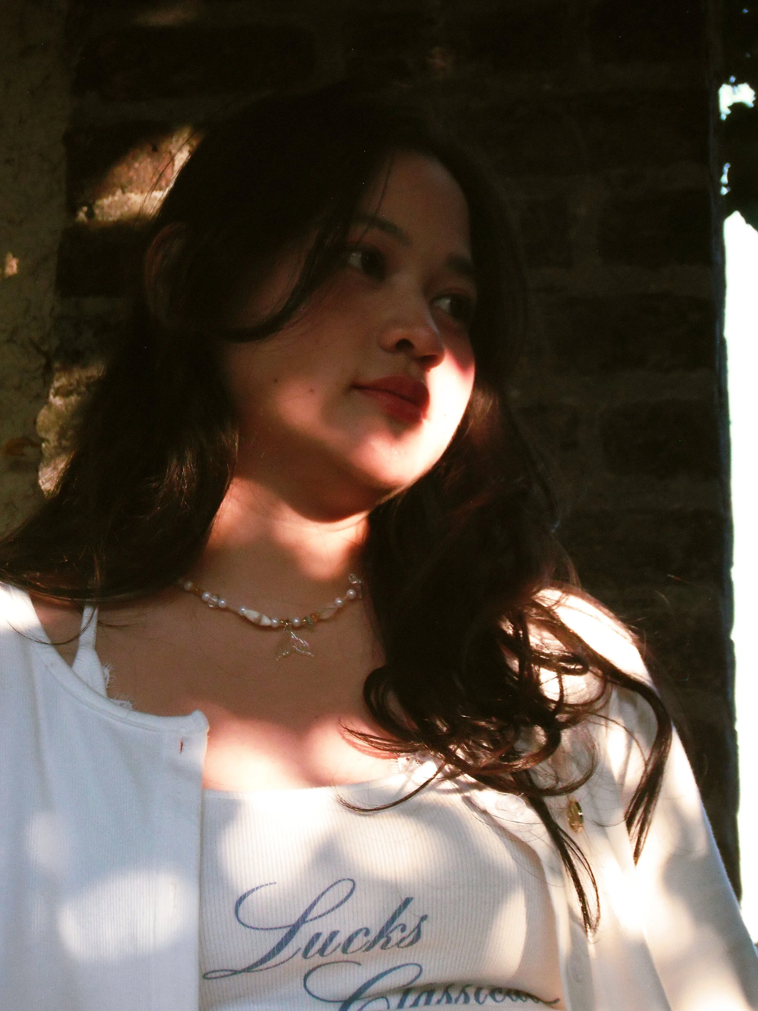 A young woman with dark hair, wearing a pearl necklace and white top with the word 'Lucks' visible, looks to the side with sunlight illuminating her face against a dark background.