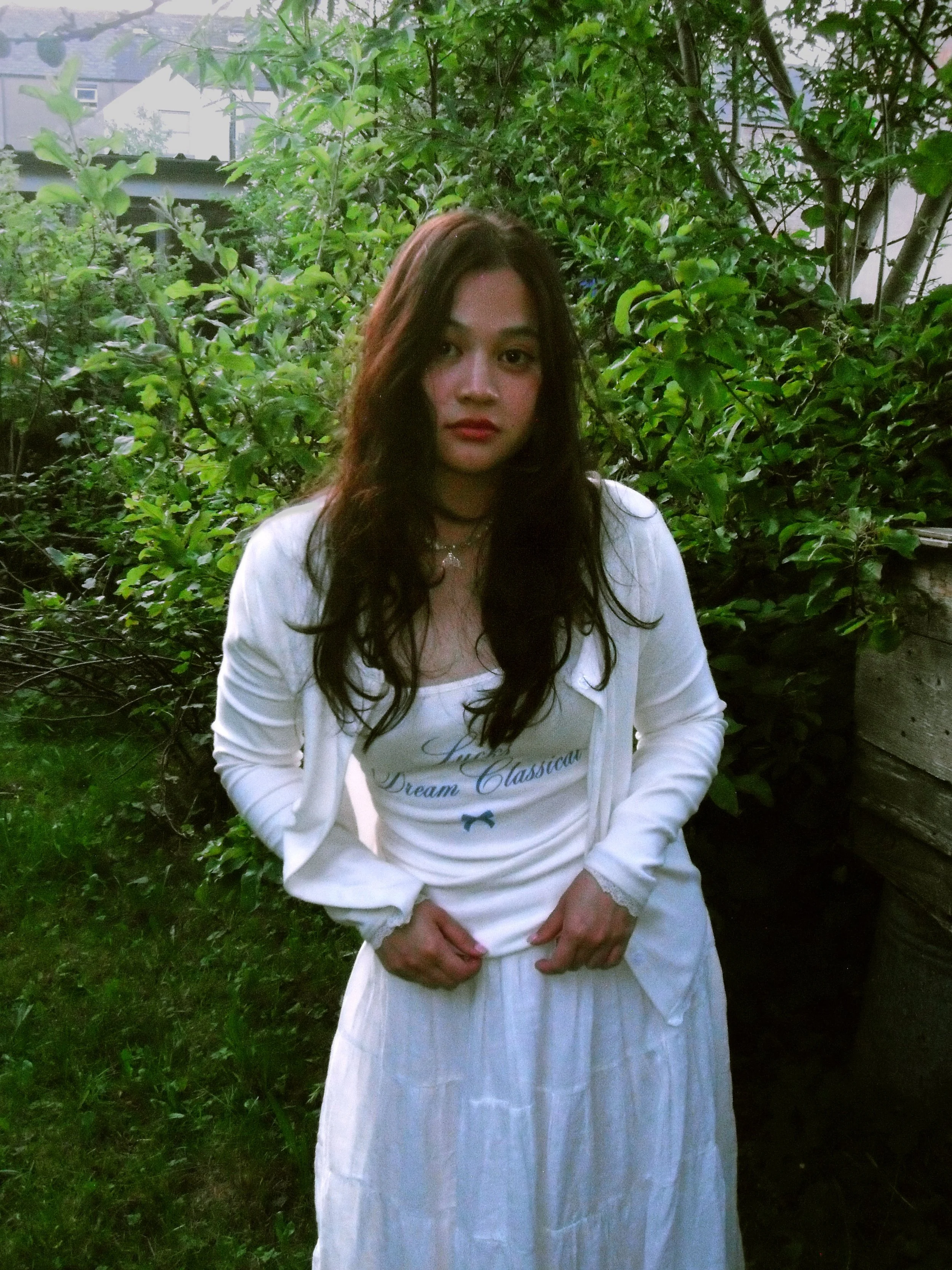 A young woman with long, dark hair standing outdoors amidst greenery, wearing a white dress and a matching white jacket, with her hands resting on her hips.