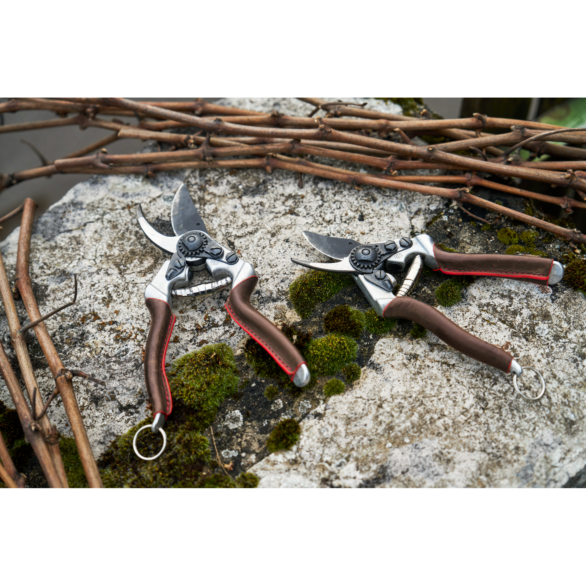 Zwei Gartenscheren mit braunen Griffen liegen auf einem moosbedeckten Stein, im Hintergrundstaket aus Zweigen.