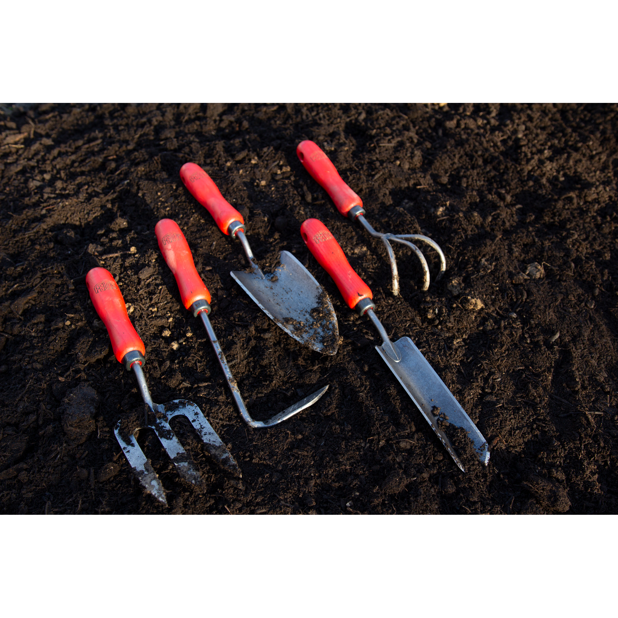 Fünf Gartenwerkzeuge mit roten Griffen liegen auf dunkler Erde.