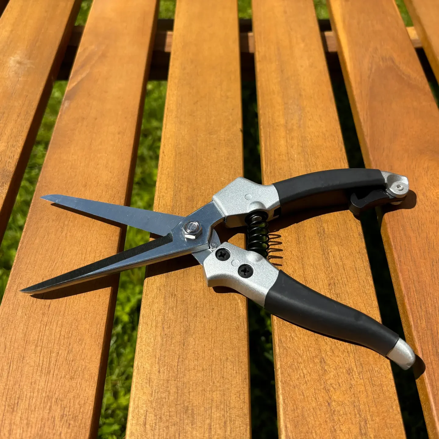 Gartenschere mit schwarzen Griffen und Metallklingen auf einem Holzbank. Im Hintergrund grüne Wiese.