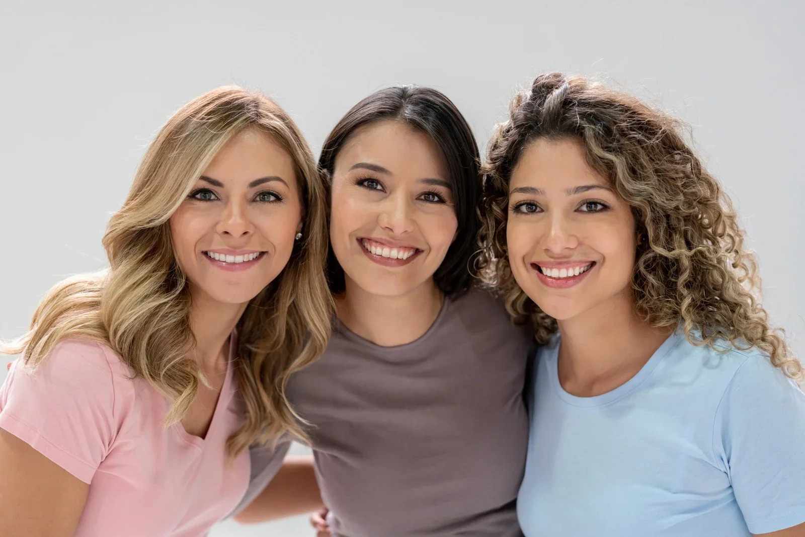 Three smiling women with different hair types and colors standing close together against a plain background.