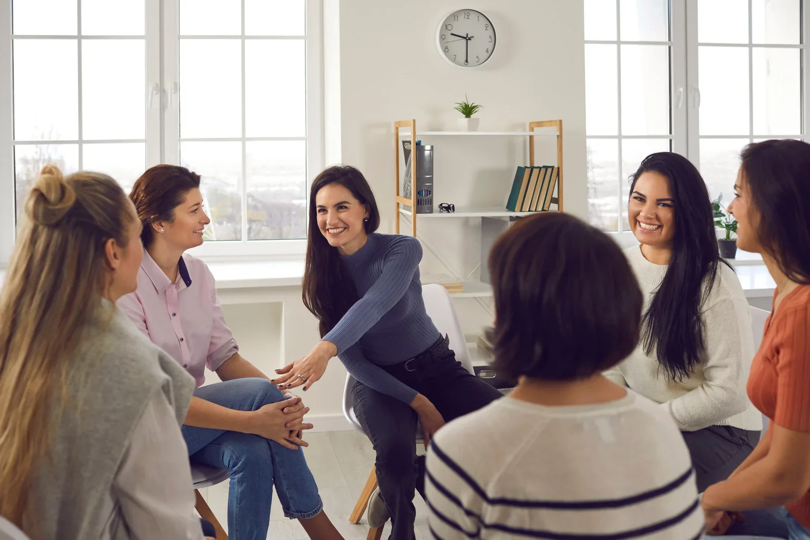 Group of women sitting in a circle in a bright, modern office, engaging in a discussion, with large windows behind them.