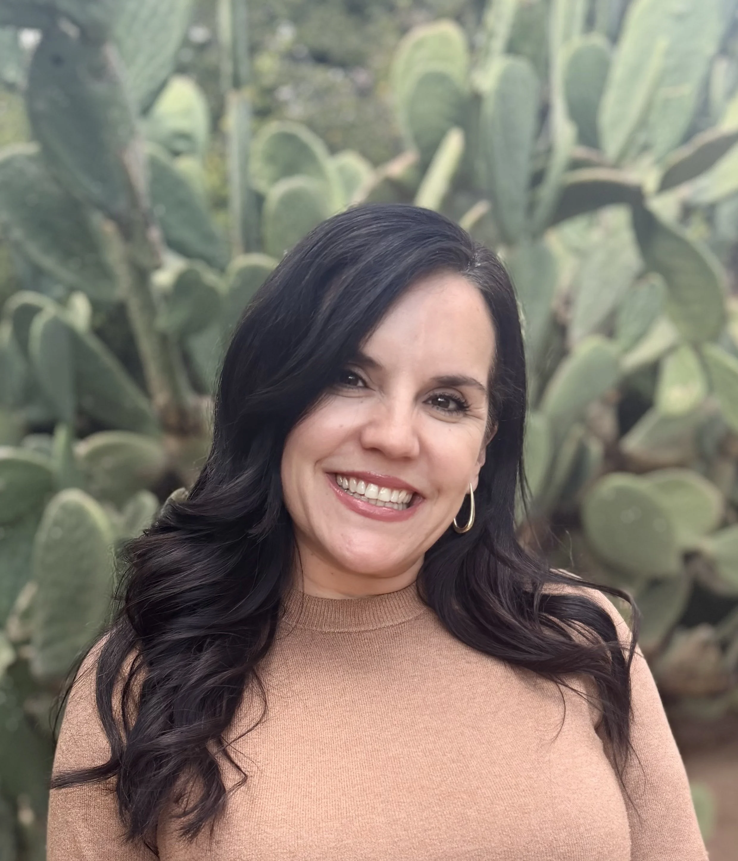 A woman with dark, wavy hair and hoop earrings smiling in front of a cactus plant.