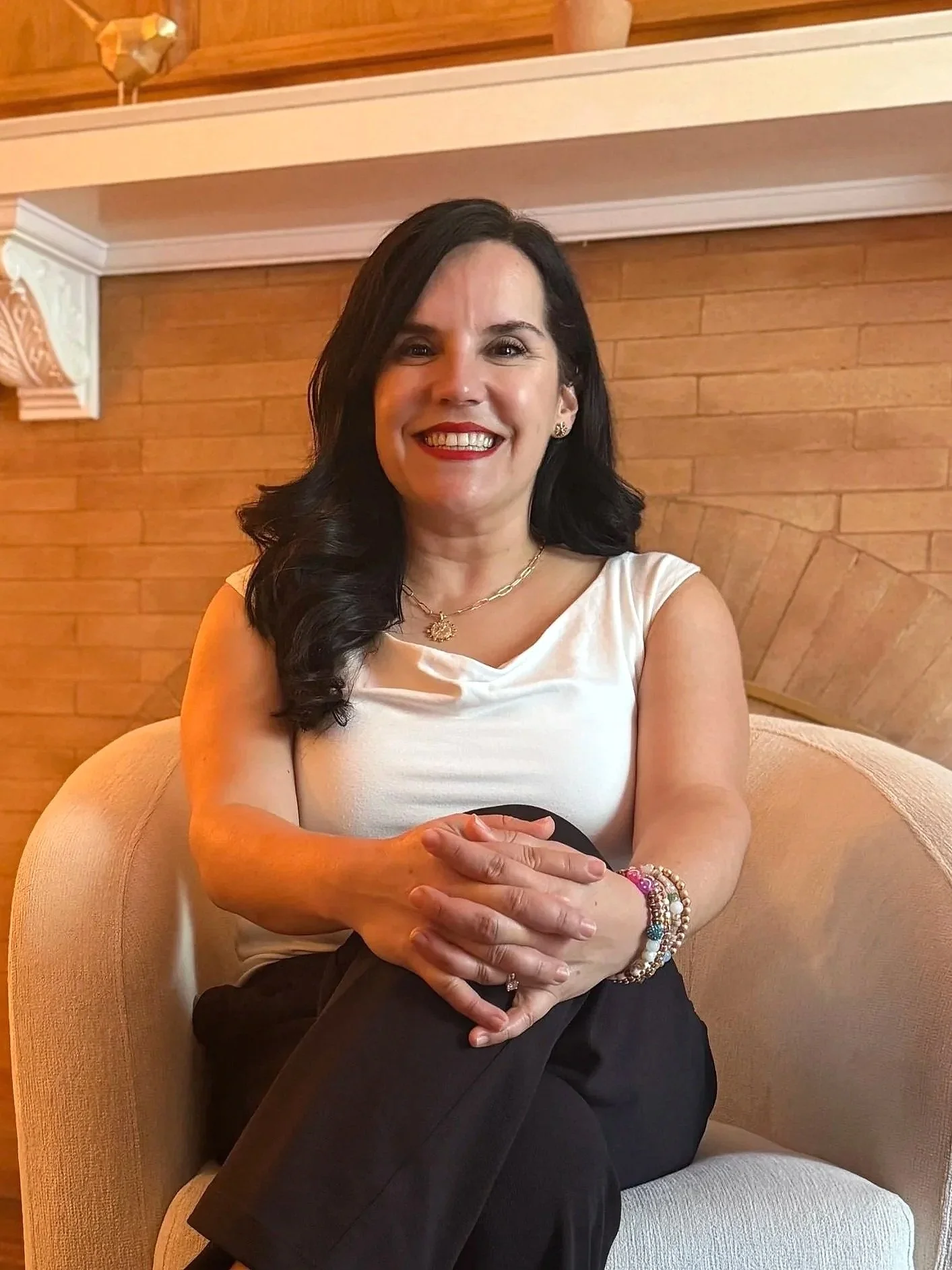 A woman with black hair, wearing a white sleeveless top and black pants, sitting on a beige armchair in front of a brick wall, smiling at the camera.