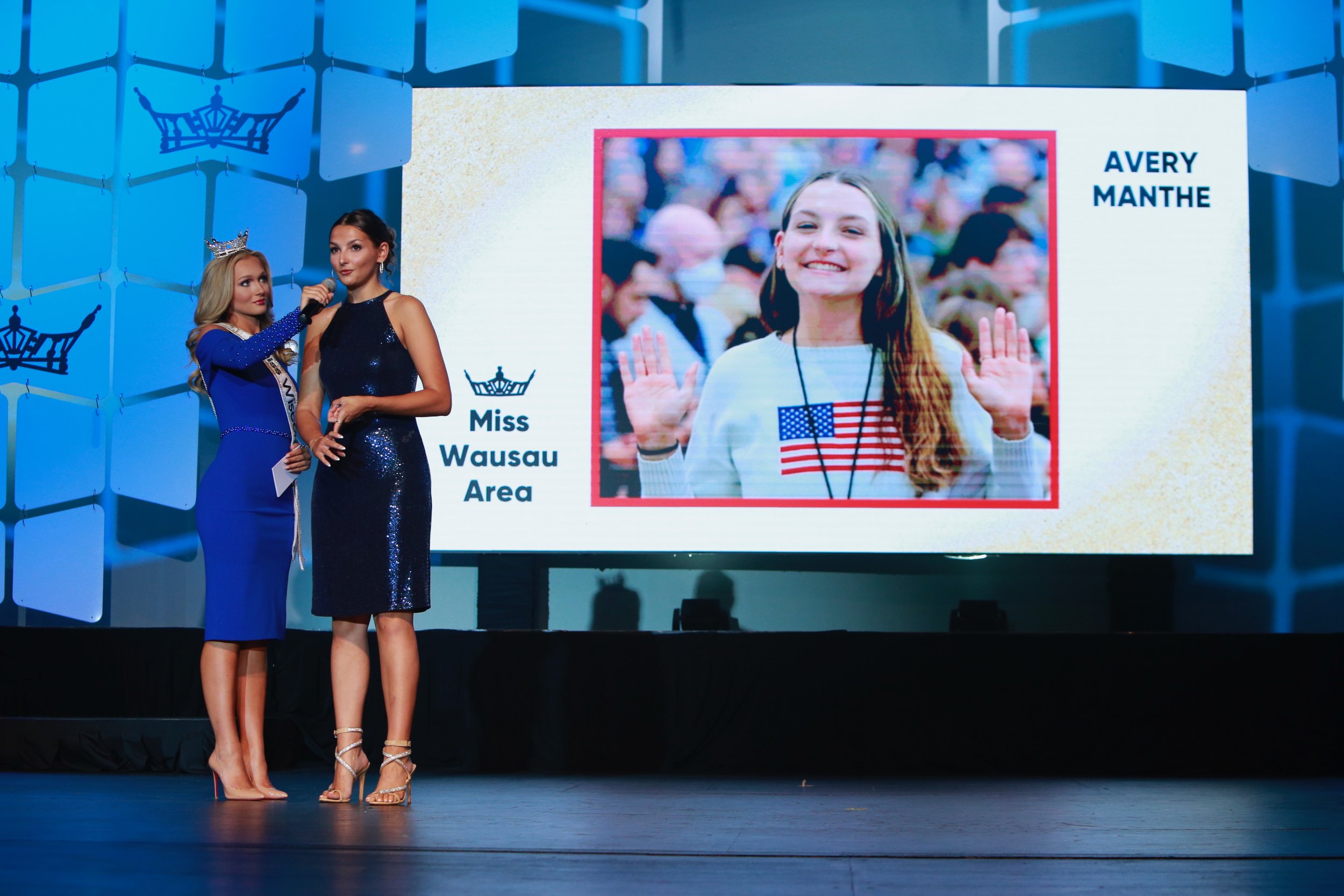Miss Wausau Area contestant being interviewed on stage during pageant event, with large screen behind showing her photo and name, Avery Manthe, in Miss Wisconsin competition.