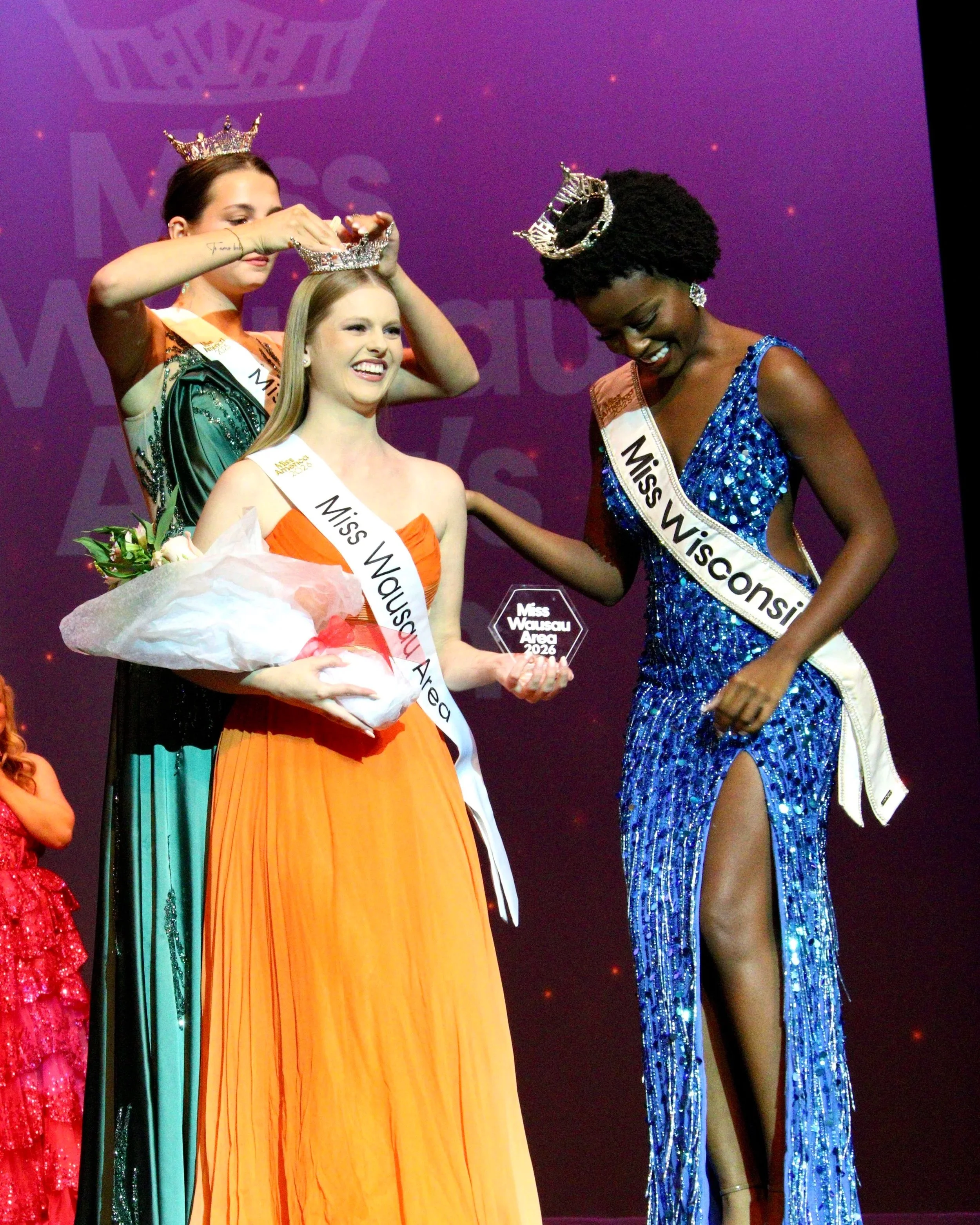 Miss Wisconsin is helping crown Miss Wausau Area 2026 on stage at a Miss Wausau Area Scholarship Competition, accepting a trophy and holding a bouquet of flowers, with Miss Wausau Area 2025, Avery Manthe, placing the crown on her head. Janae Bond.