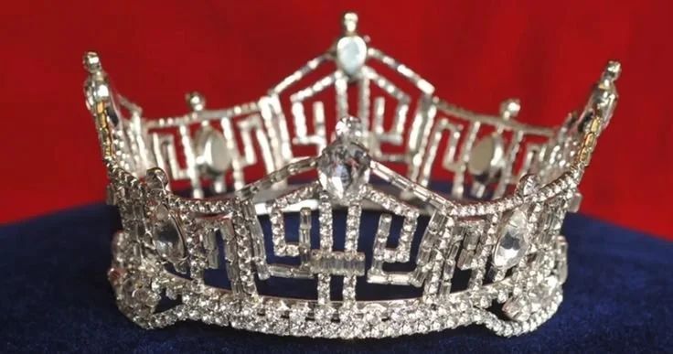 A silver tiara adorned with numerous clear gemstones, set against a red background with a dark surface below. Miss America crown.