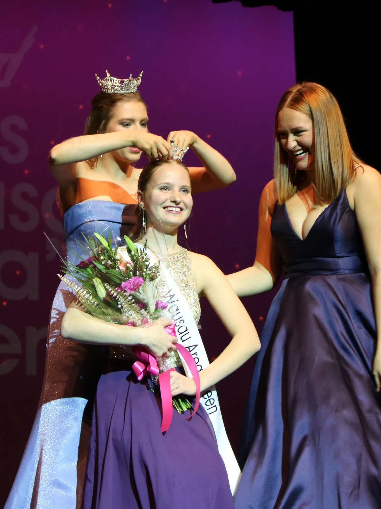 A young woman being crowned as winner on stage, holding a bouquet of flowers, with two women beside her helping or celebrating. Miss Wausau Area's Teen 2025 Anna Jacobson is crowned as the winner of the Miss Wausau Area Scholarship Competition.