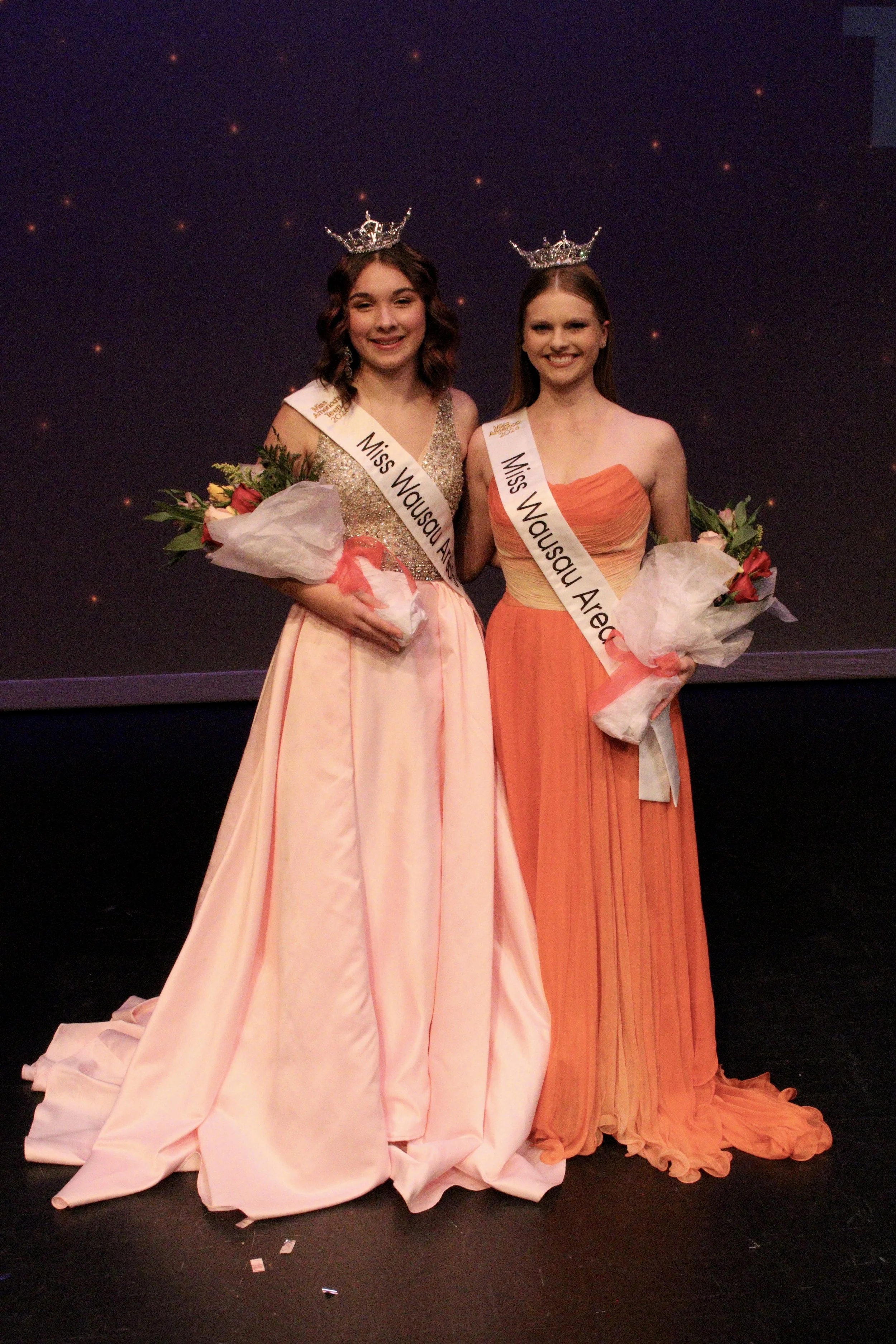 Two young women wearing coronets and sashes that read 'Miss Wausau Area' are holding bouquets of flowers and posing together on stage, smiling at the camera. Miss Wausau Area's Teen. Chloe Munguia, Janae Bond.
