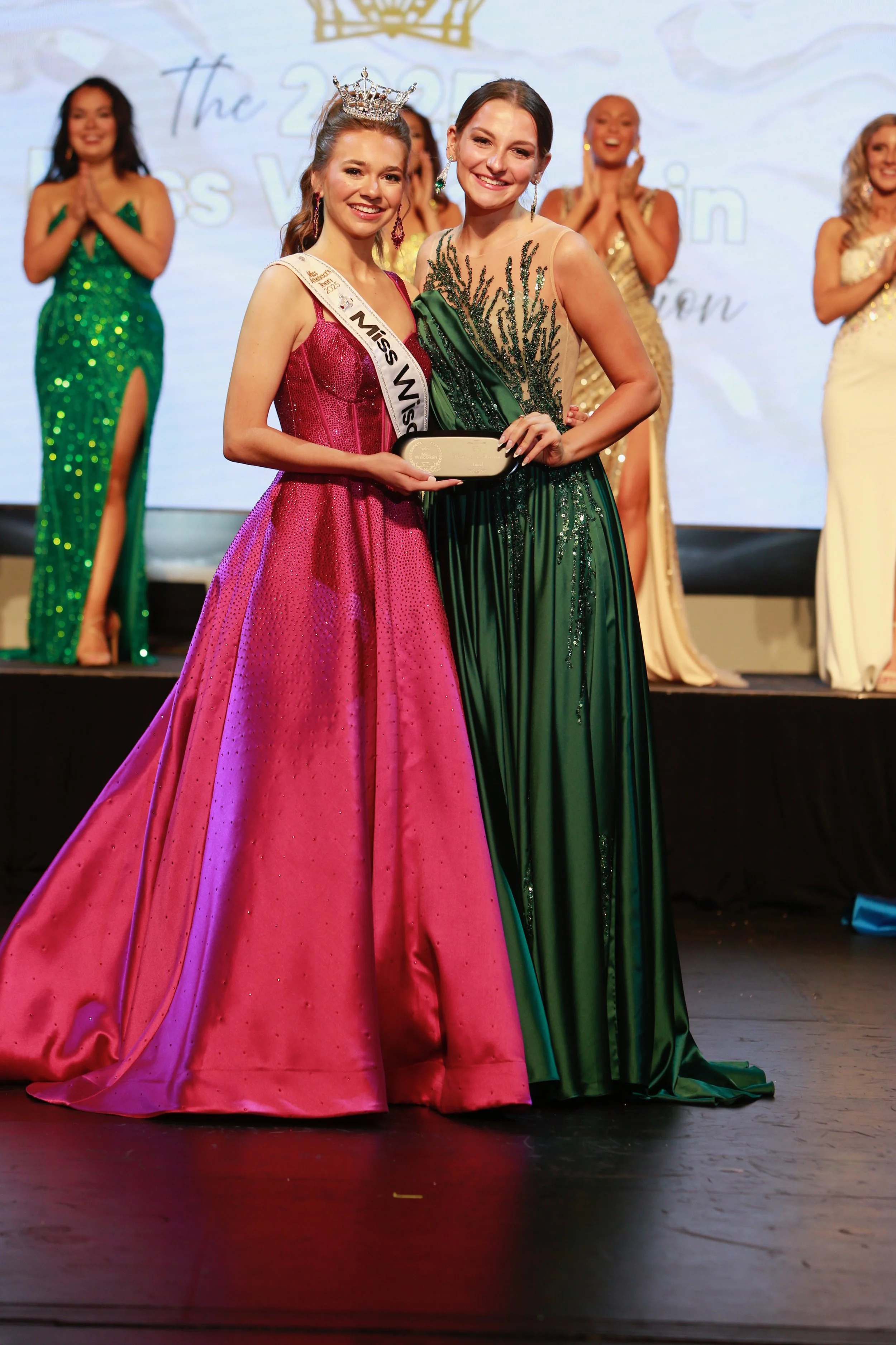 Miss Wisconsin's Teen wearing a pink ball gown and sash, providing an award to Miss Wausau Area 2025, Avery Manthe, in a green gown during a beauty pageant. Several other women in gowns watch and applaud in the background.