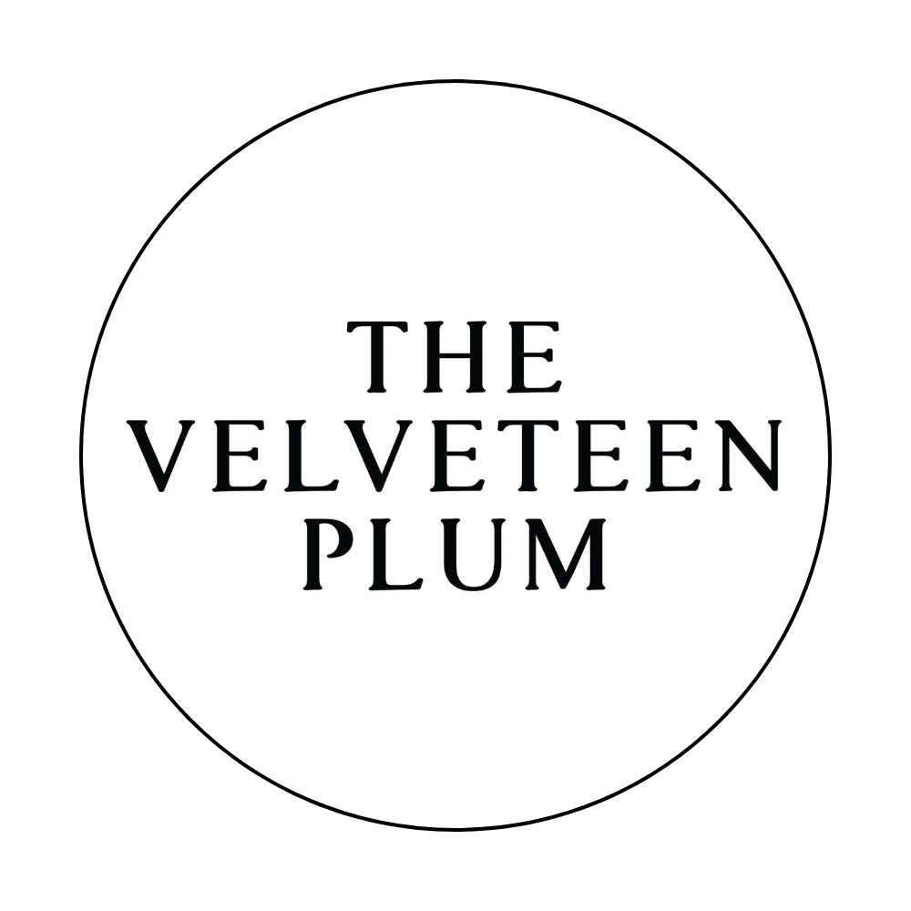 Logo with black text that reads 'The Velveteen Plum' inside a thin black circle on a white background.