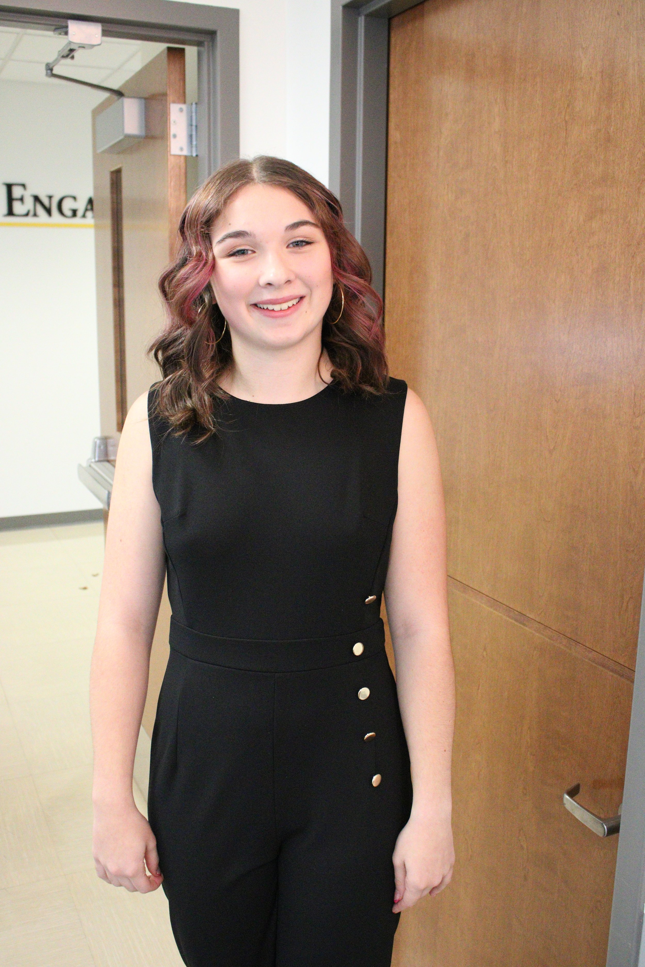 Young woman with wavy, shoulder-length hair and hoop earrings, smiling and standing in front of a wooden door in an indoor setting. Miss Wausau Area's Teen 2026 Chloe Munguia