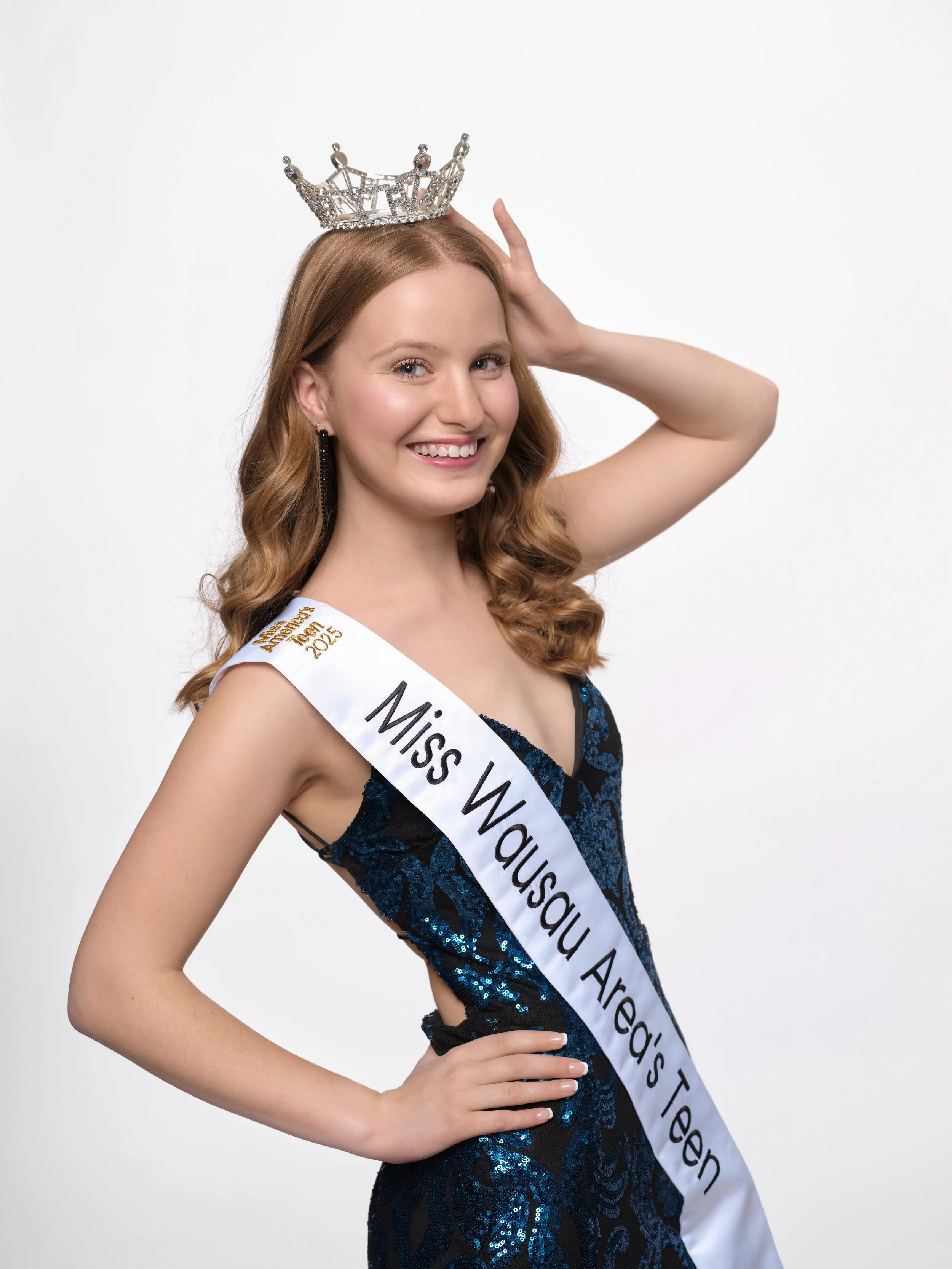 A young woman wearing a crown, sash, and a black and blue dress, smiling and posing against a plain white background.