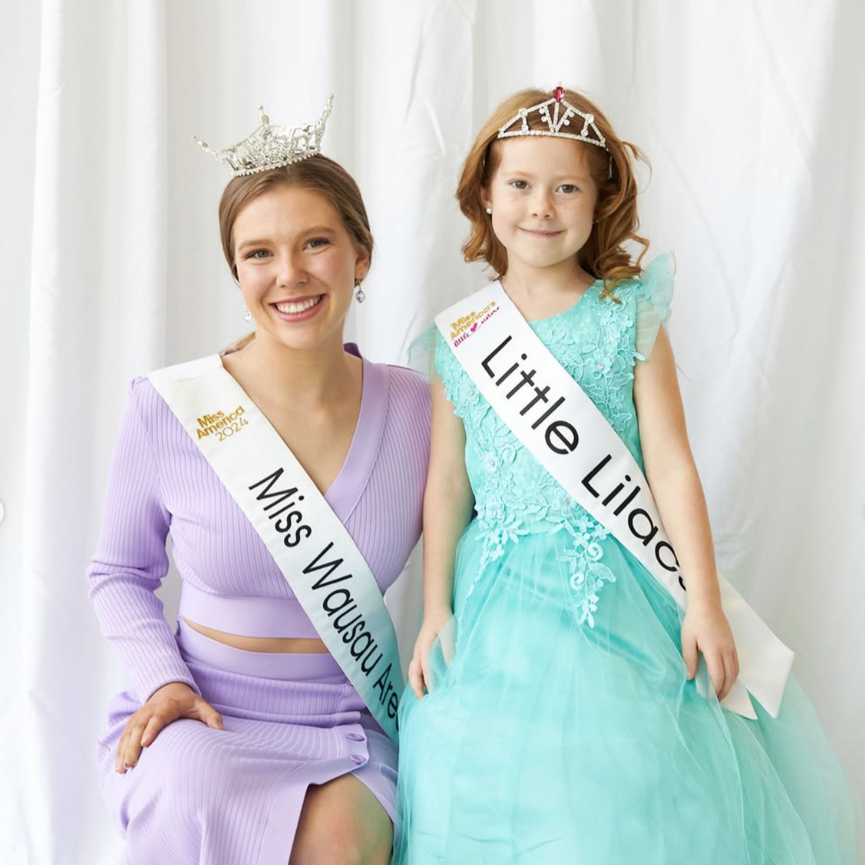A woman and young girl sitting together, both wearing tiaras and sashes, smiling at the camera against a white background. The woman is Miss Wausau Area 2024, Lauren wearing a lavender dress, and the girl is Little Lilac, dressed in a turquoise gown.