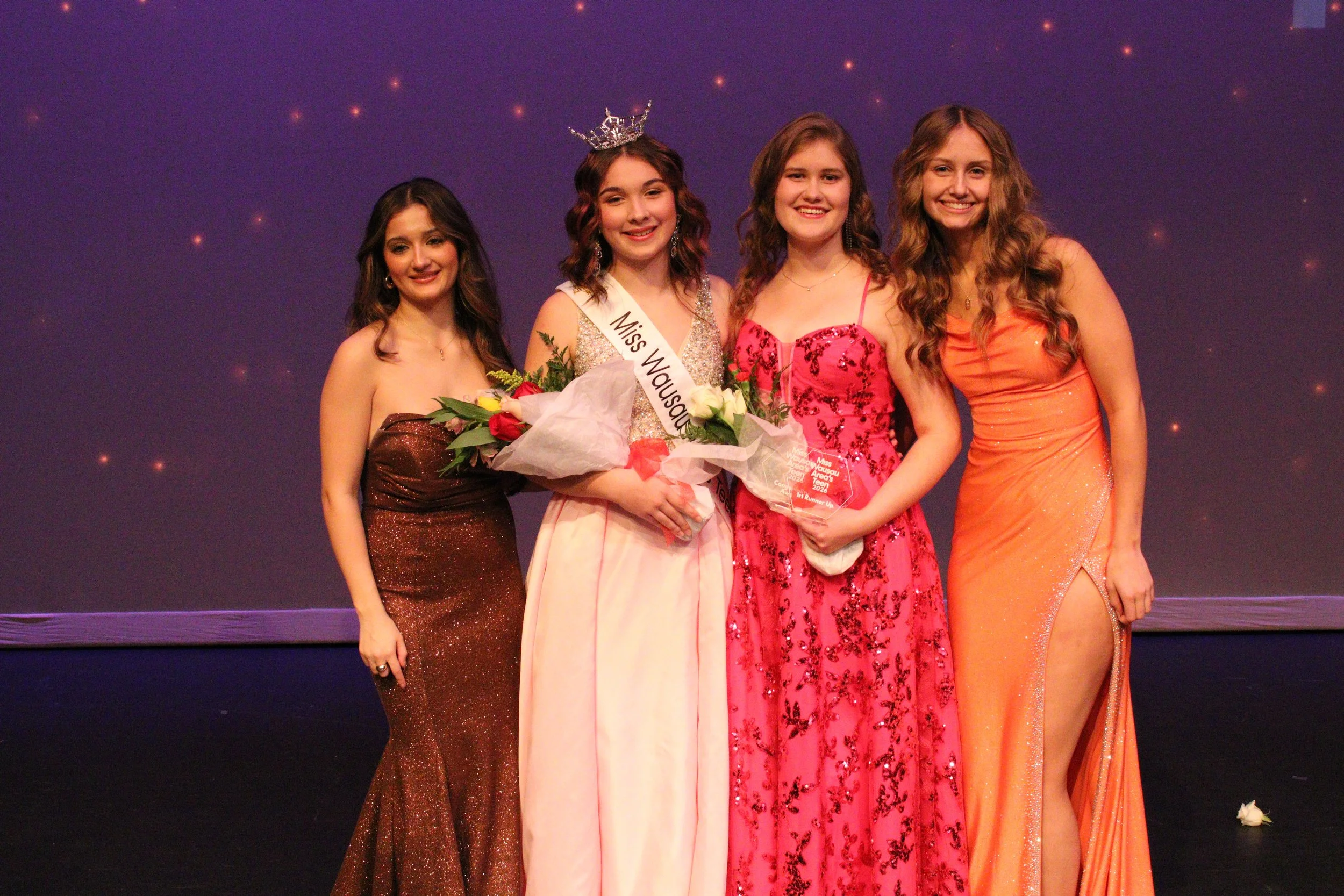 Group of four women standing on stage during a beauty pageant, with one woman holding a bouquet of flowers and wearing a crown, others in elegant evening dresses. Miss Wausau Area's Teen 2026 class of contestants.