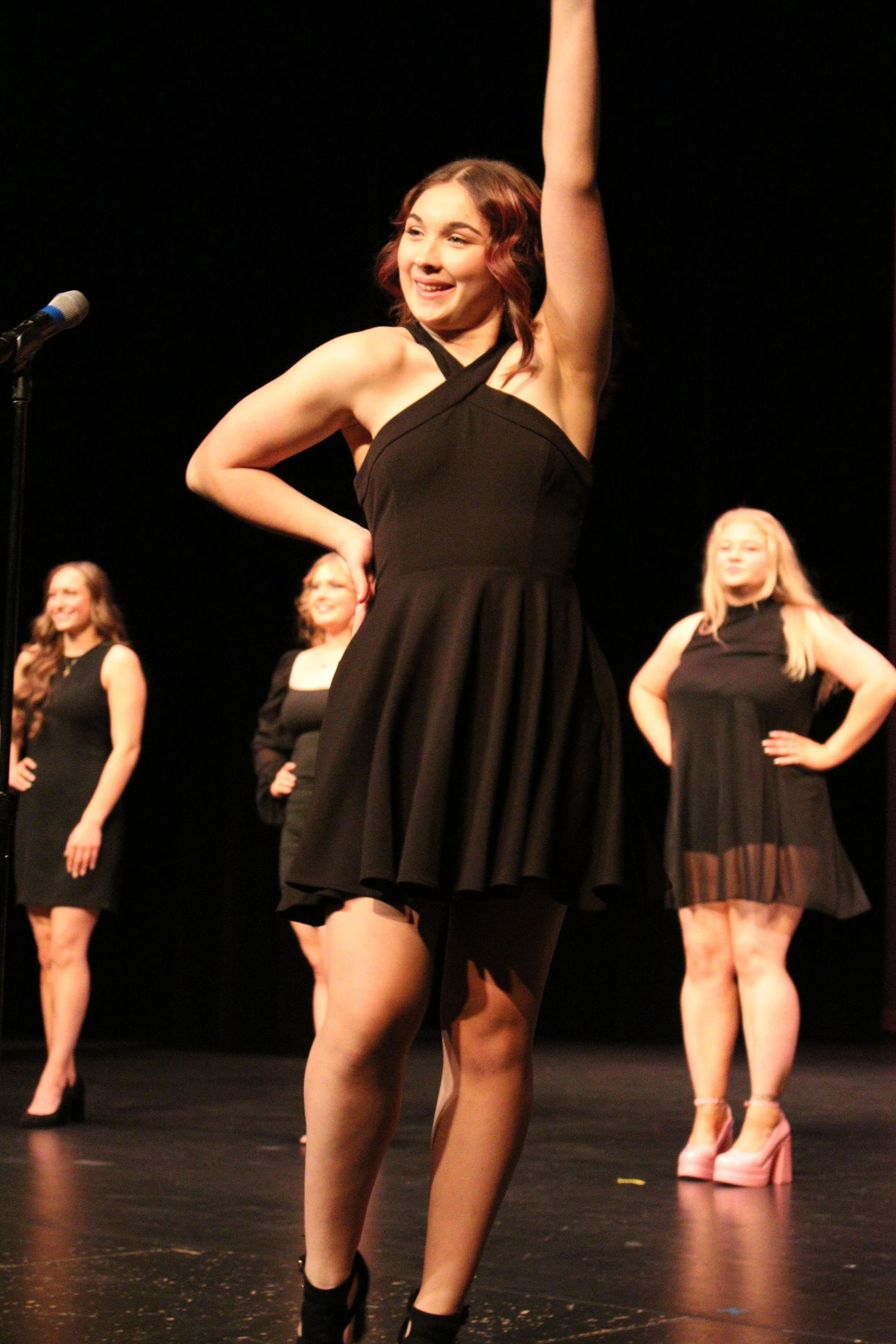 A young woman in a black dress with a halter neckline poses on stage with her right arm raised and her left hand on her hip, smiling. Three other women in black outfits and high heels stand behind her on a dark stage. Miss Wausau Area's Teen