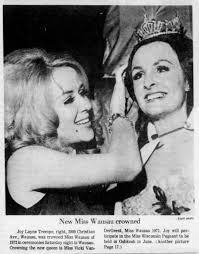 Two women smiling, one wearing a crown and sash, the other placing a crown on her head, at a beauty pageant.