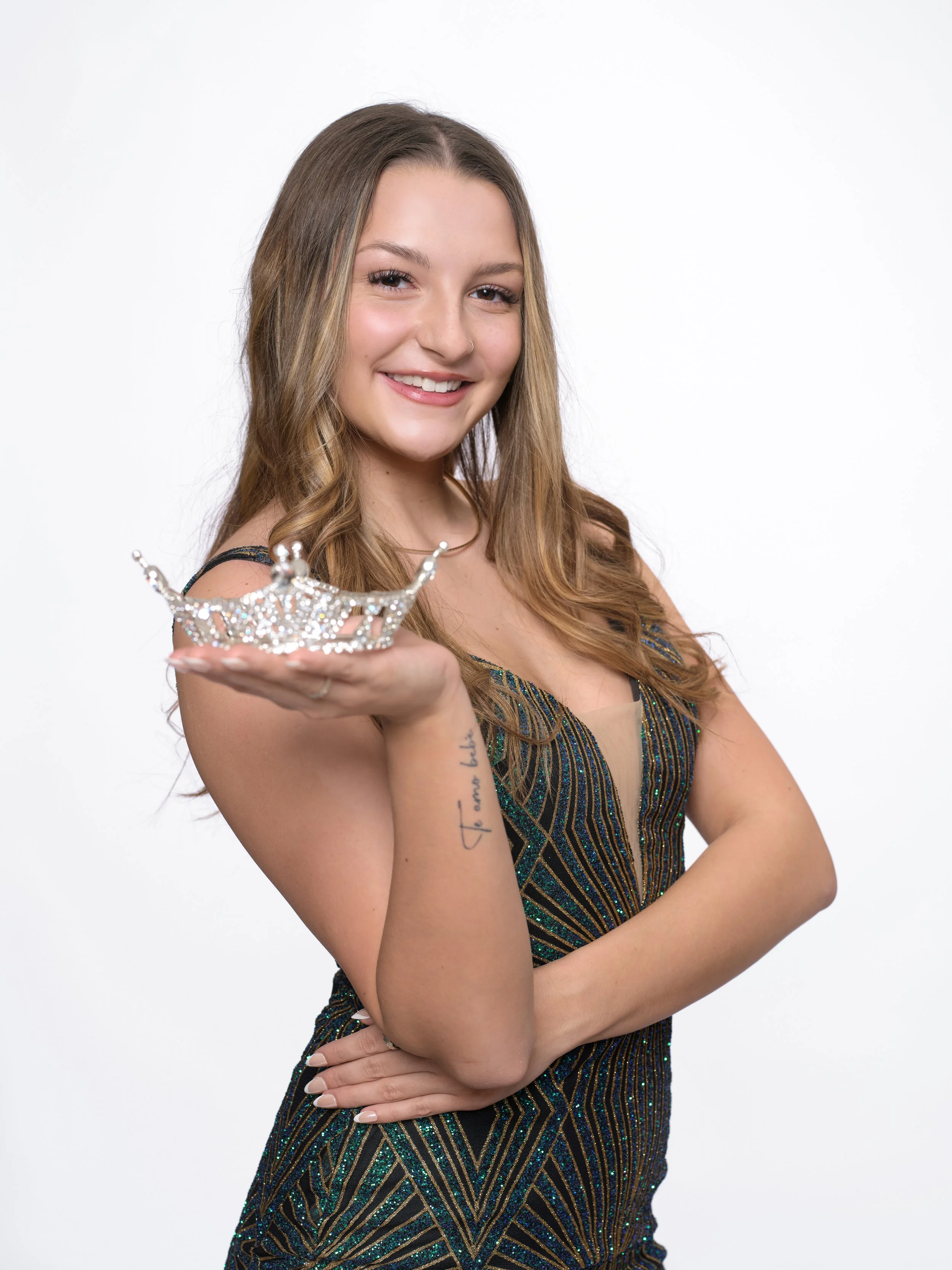 A young woman with long, wavy hair, wearing a sparkly, patterned dress, holding a small tiara in her right hand, smiling at the camera, with a white background.