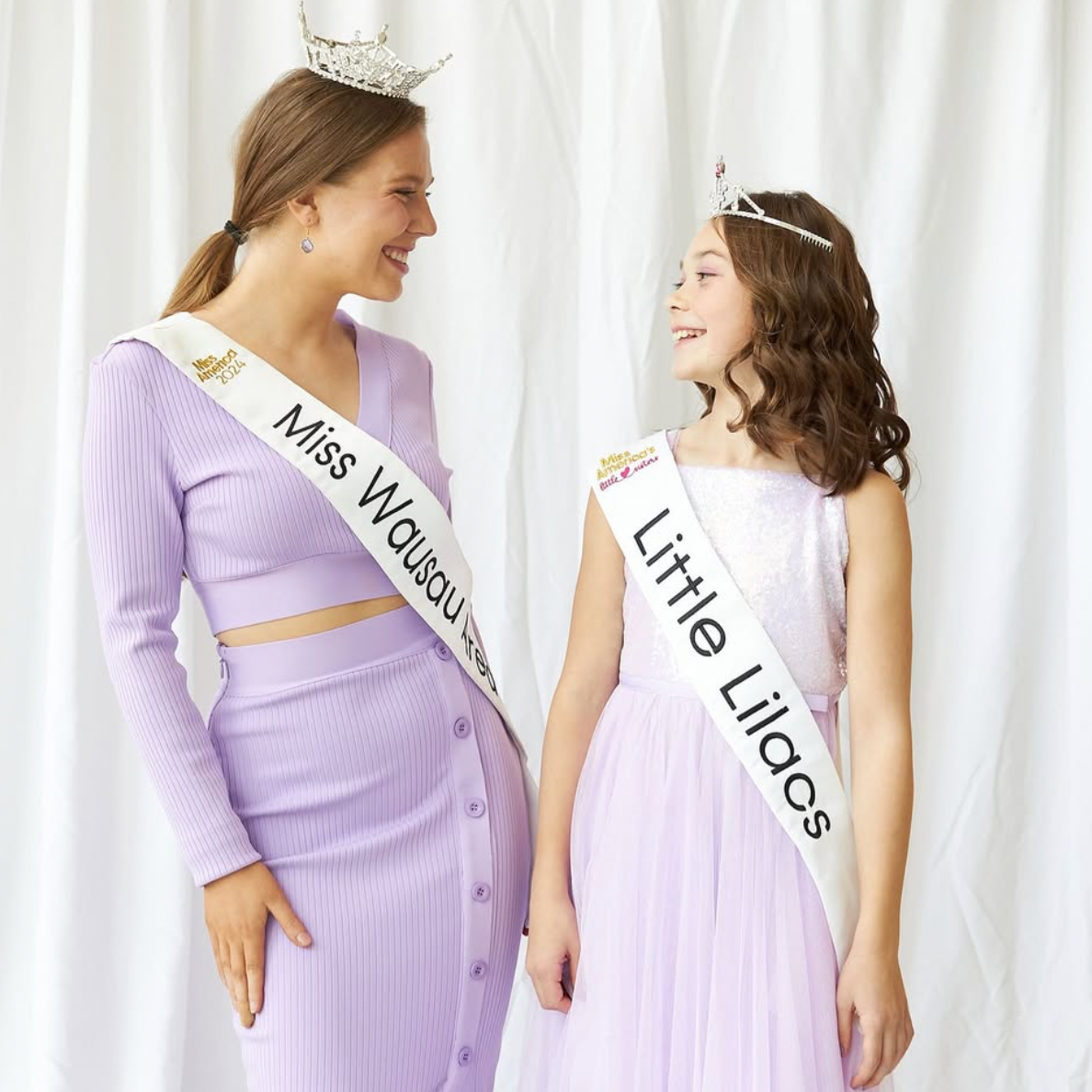 Two women wearing tiaras and sashes; the woman on the left is Miss Wausau Area 2025, Lauren Hjelsand, and the woman on the right is Little Lillacs, both smiling and looking at each other, standing in front of a white curtain.