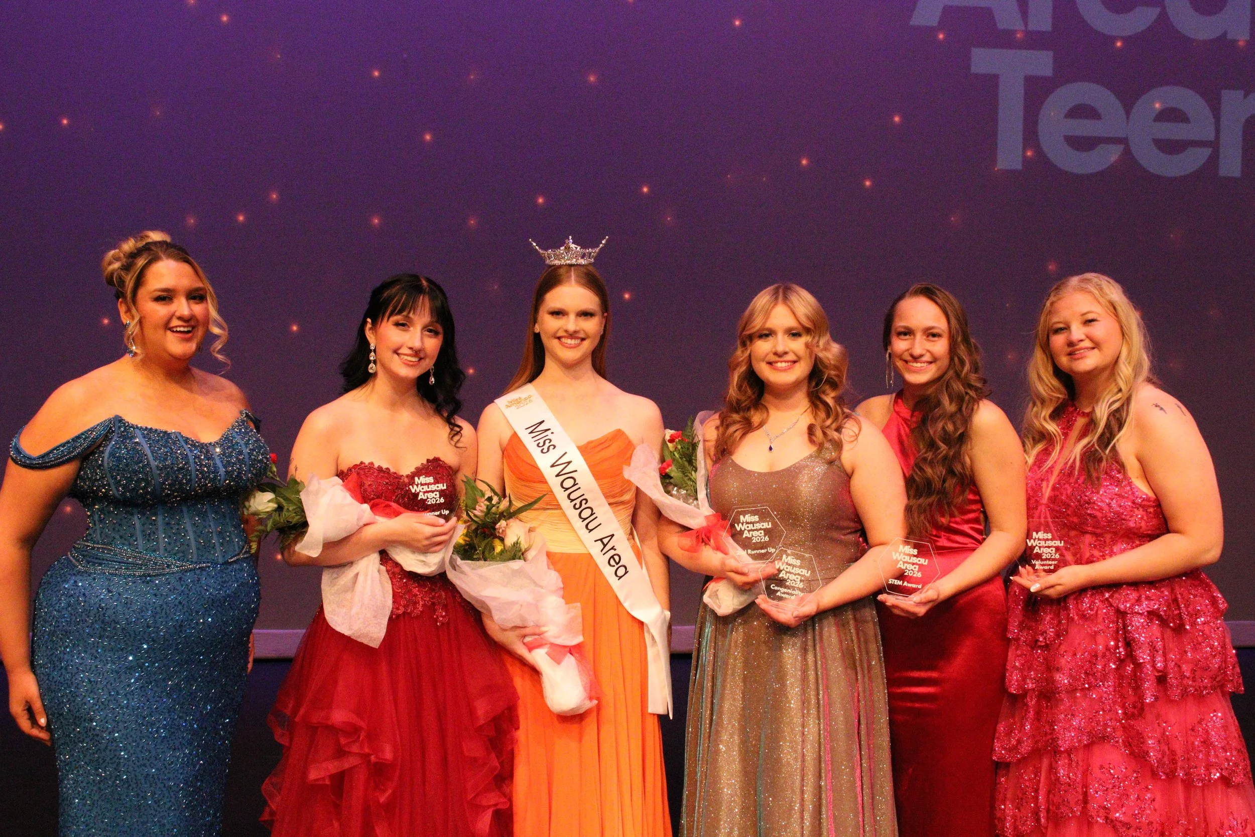 Six young women dressed in evening gowns standing on stage with awards and flowers, one in the center wearing a sash and crown, at a beauty pageant event. Miss Wausau Area 2026 class of contestants.