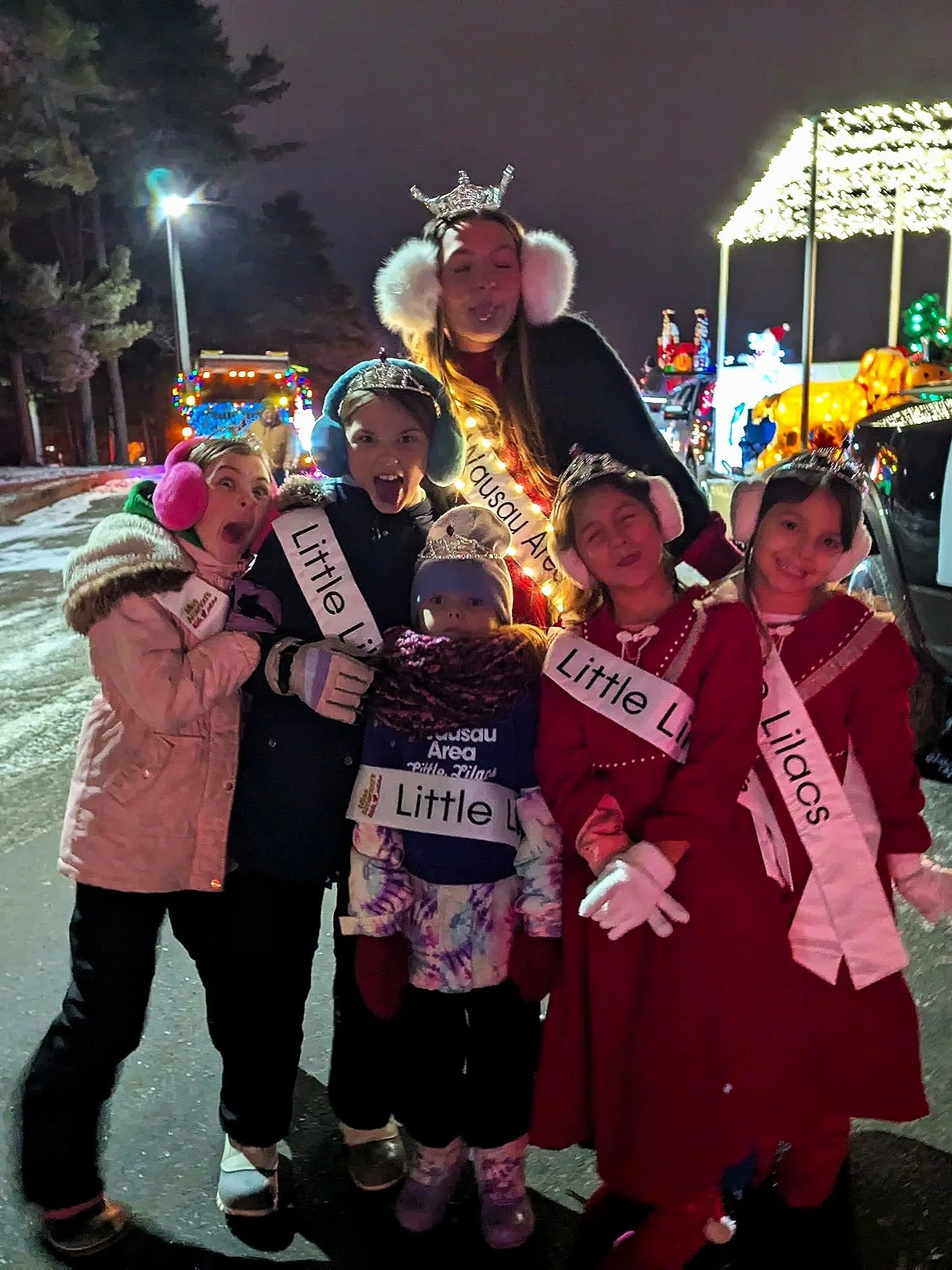 Group of children and Miss Wausau Area 2025, Avery Manthe at a nighttime holiday parade, wearing earmuffs and sashes that say 'Little Licas,' standing in front of decorated floats.