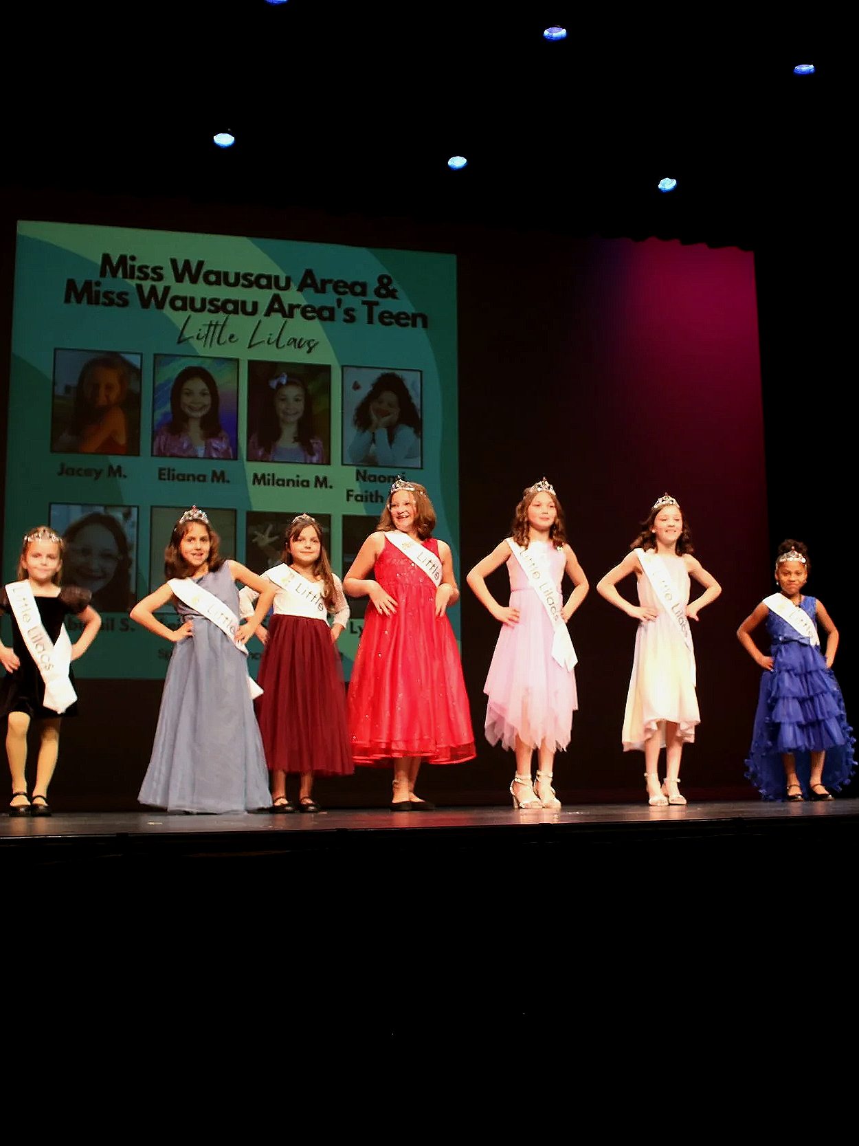 Six young girls wearing elegant dresses and tiaras on stage during a beauty pageant, with a large screen behind them displaying their names and photos.