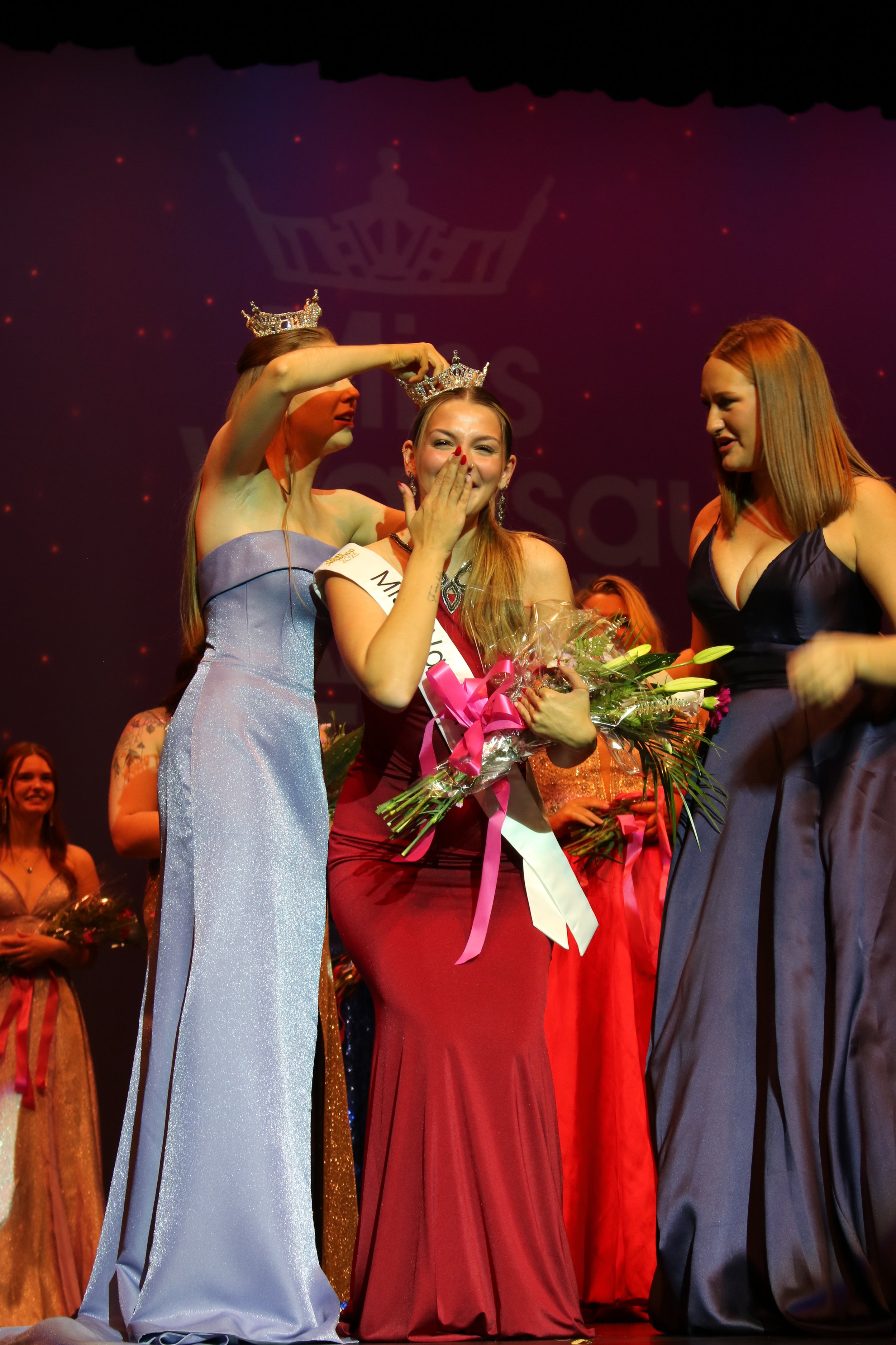 A beauty pageant winner with a sash, crowned, and holding a bouquet of flowers, being crowned by another contestant on stage, during the pageant's moment of celebration. Avery Manthe crowning moment.