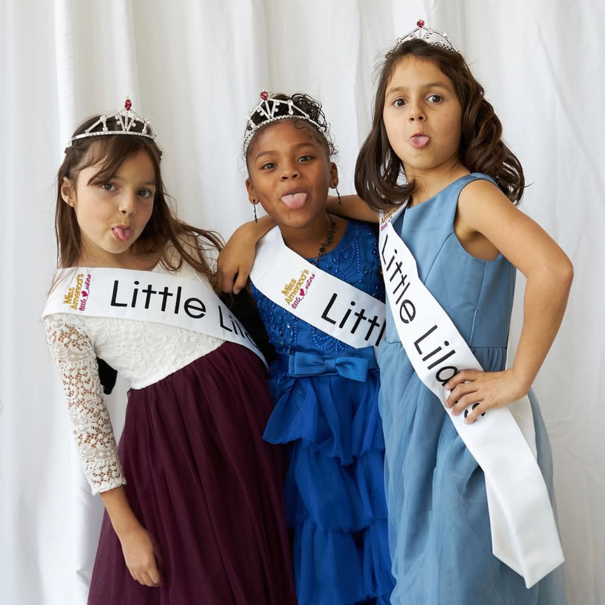 Three young girls wearing princess dresses and tiaras, making silly faces and sticking out their tongues. They have sashes that say 'Little Lilacs' and are smiling happily, with a white curtain as the background.