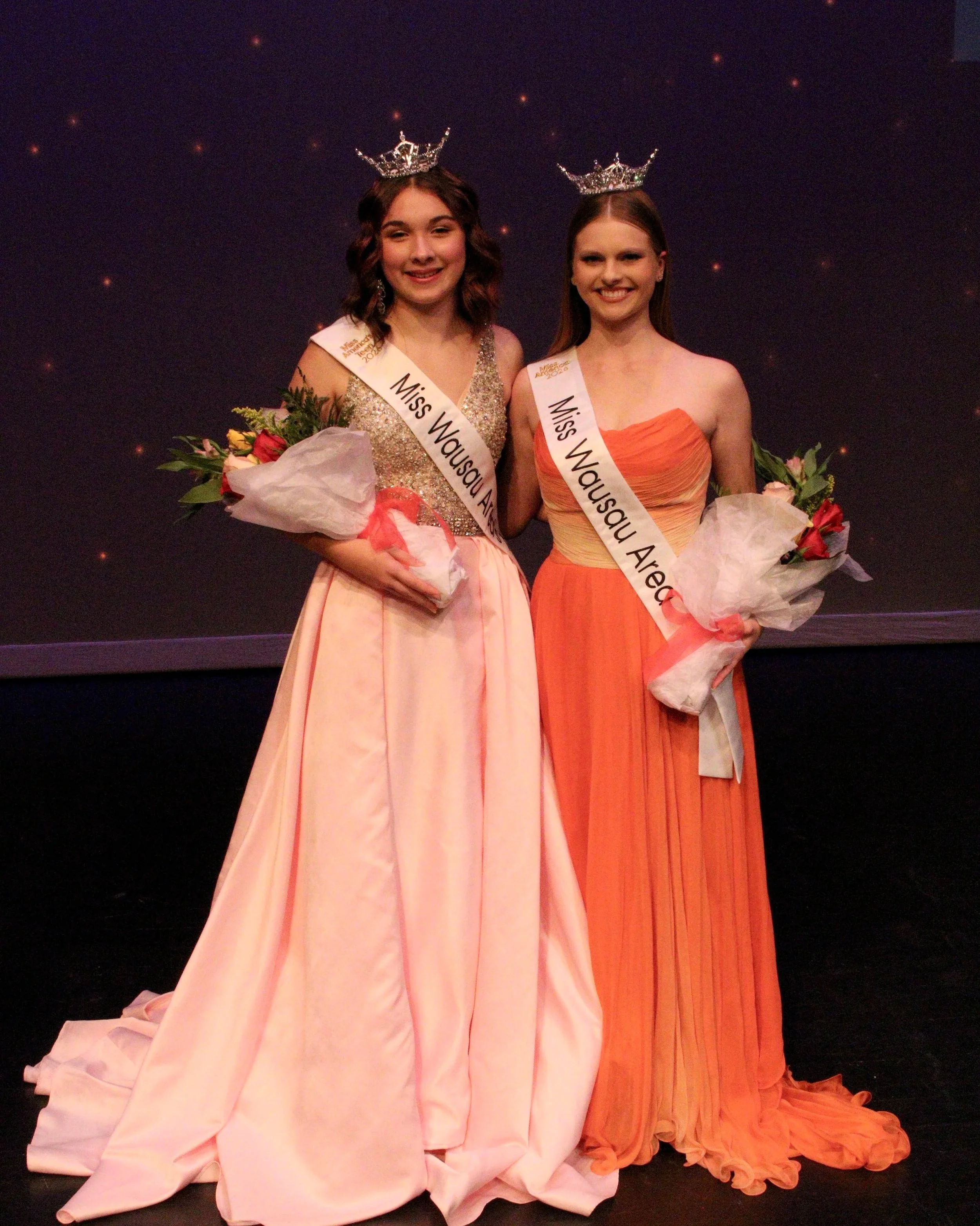 Two young women wearing crowns and sashes that read 'Miss Wausau' and 'Miss Wausau Area' stand together, each holding a bouquet of flowers wrapped in white tissue paper, on a dark background with small lights. Janae Bond and Chloe Munguia.