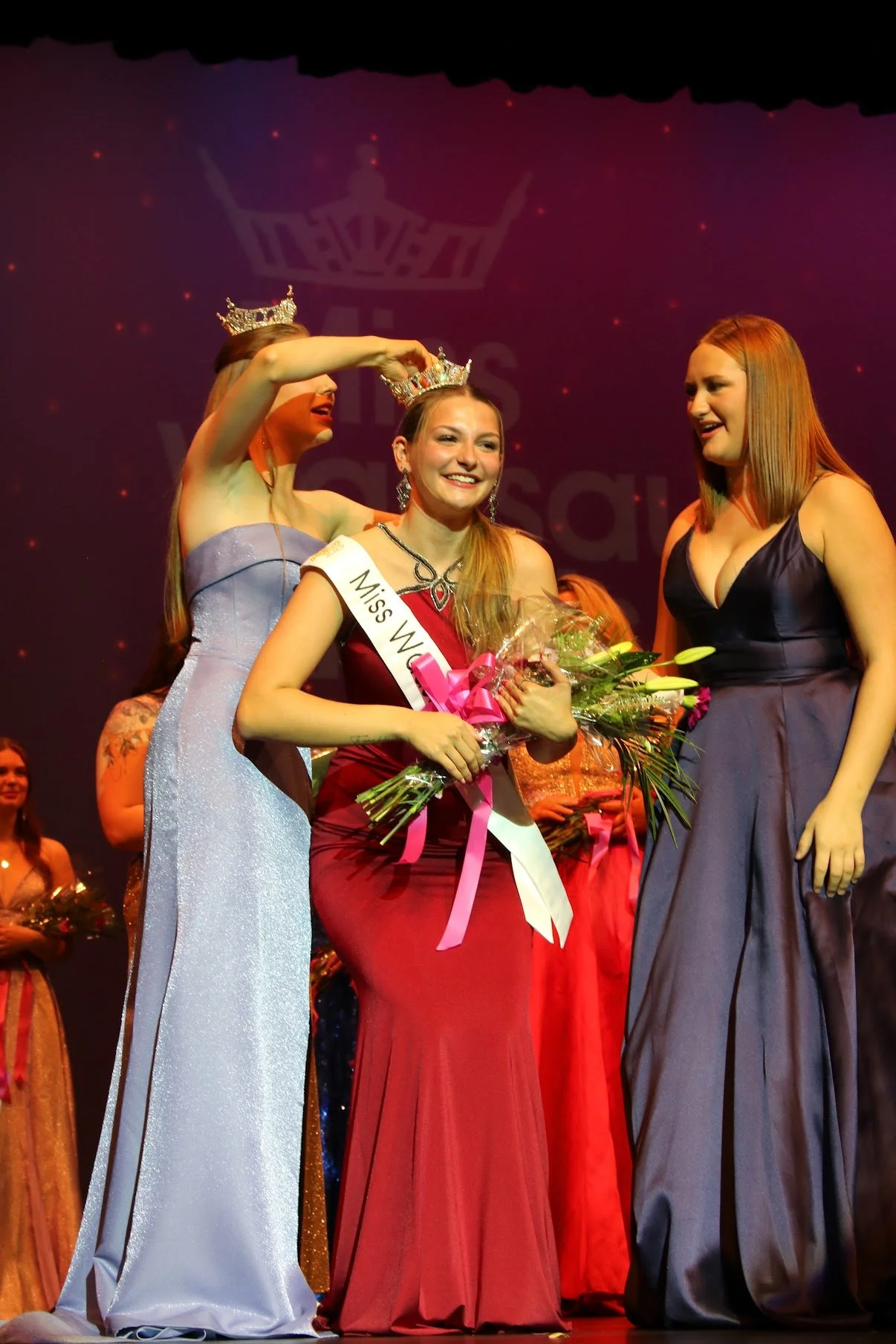 Miss Wisconsin wearing a crown and sash, holding flowers, being crowned during a beauty pageant stage with other contestants in the background. Miss Wausau Area 2025 Avery Manthe is crowned the winner of the Miss Wausau Area Scholarship Competition.