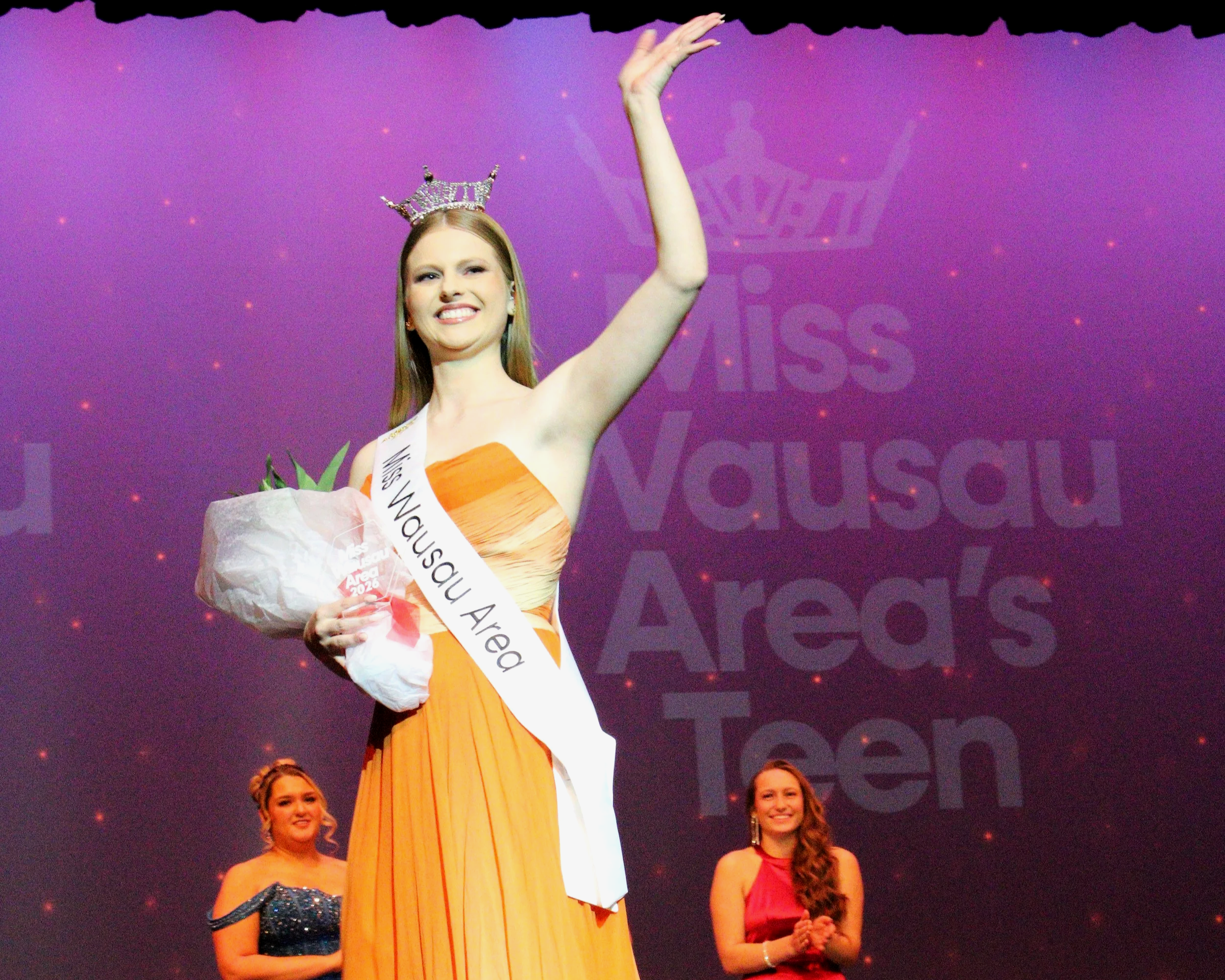 Miss Wausau Area 2026 wearing a crown and sash, holding a bouquet of flowers, on stage at the Miss Wausau Area Scholarship Competition, with women clapping in the background. Janae Bond.