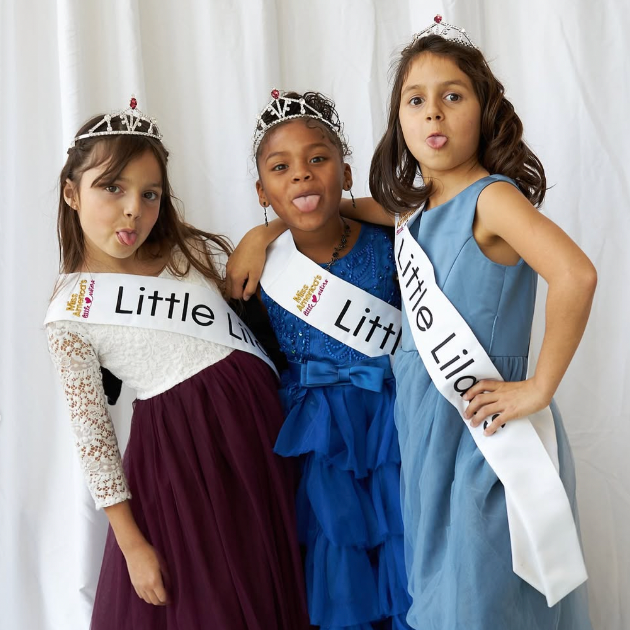 Three young girls wearing tiaras and sashes that read 'Little Lilacs' pose with playful expressions, sticking out their tongues in front of a white backdrop.