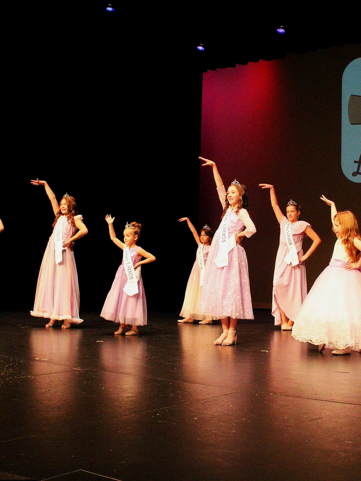 Young girls wearing tutus and tiaras, participating in a beauty pageant on stage, with sashes that read 'Little Lilacs'.