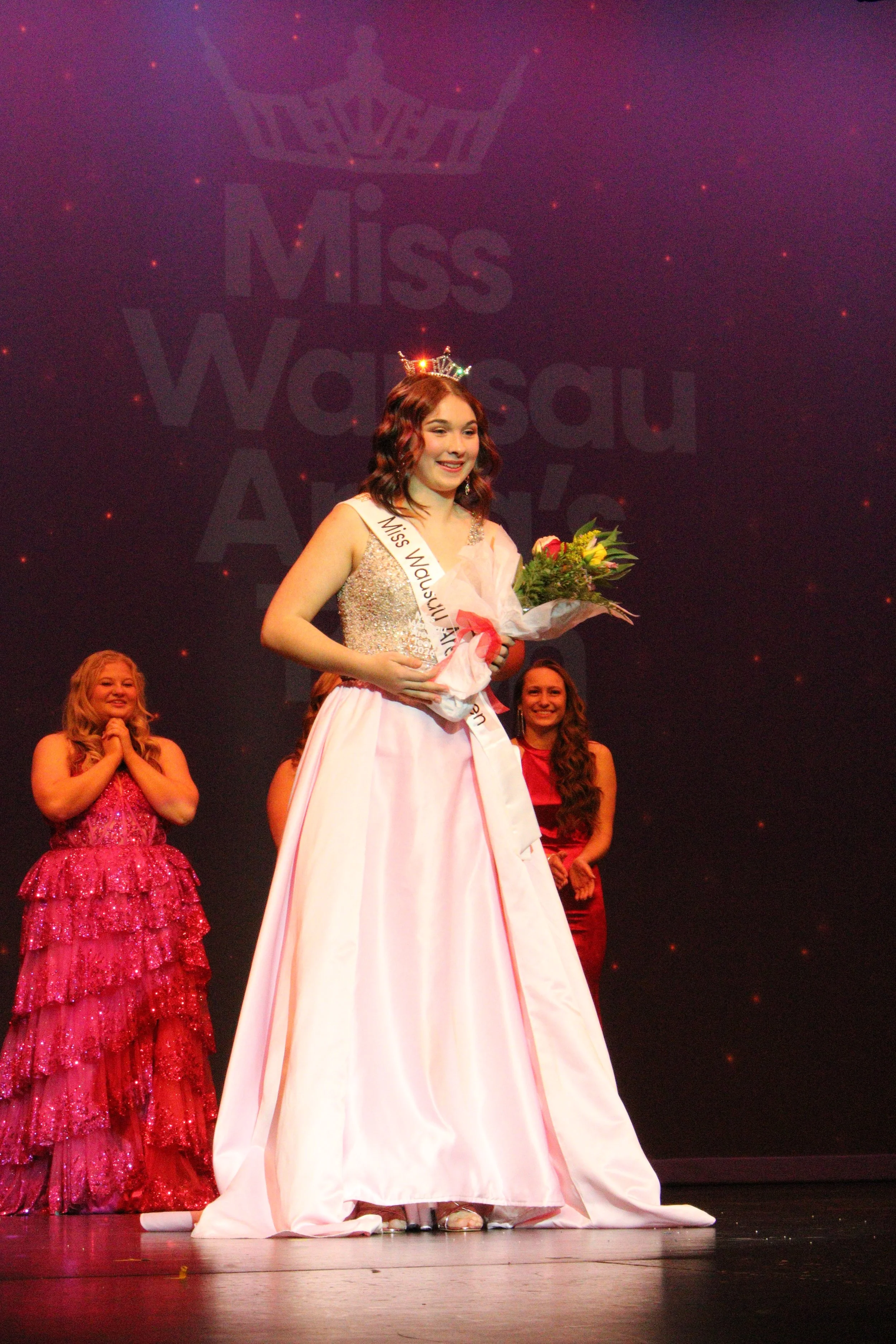 Miss Wausau Area's Teen 2026, Chloe Munguia, winner on stage, wearing a crown, holding a bouquet of flowers, in a pink gown, smiling, with contestants in dresses behind her.