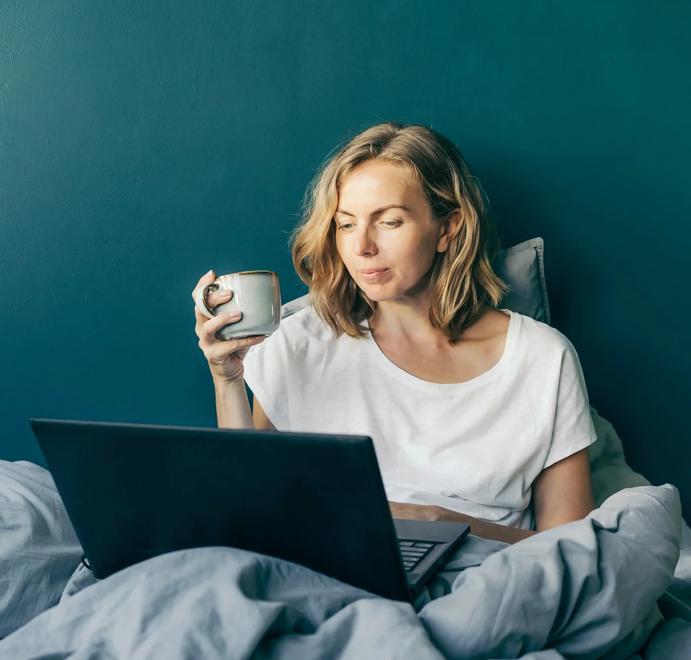 A person sitting in bed using a laptop and holding a mug during a virtual fertility therapy session.
