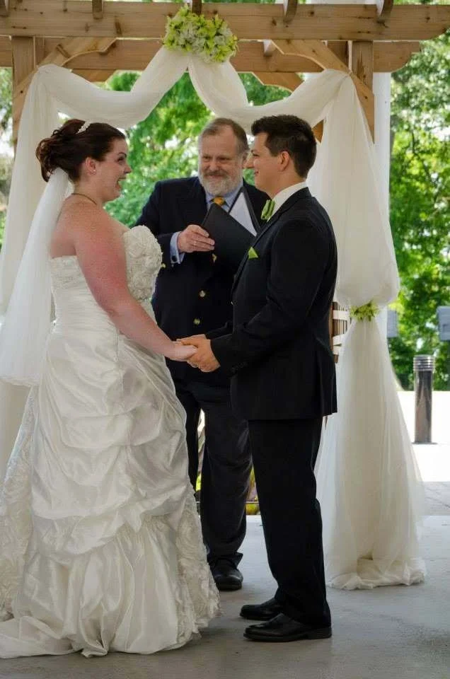 Ashley and her husband laughing and holding hands during their wedding ceremony under a floral arbor