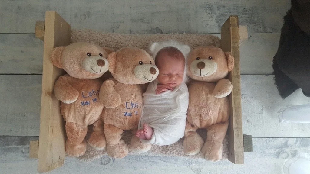 A newborn swaddled between three remembrance teddy bears embroidered with their names and dates