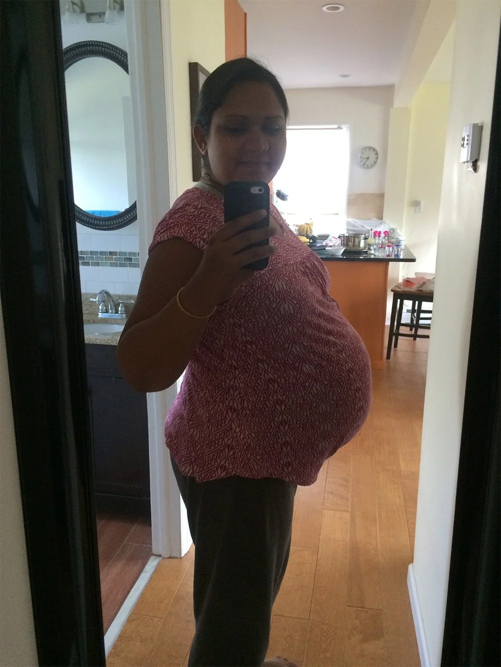 Rina taking a mirror selfie in a hallway, visibly pregnant, wearing a patterned top.