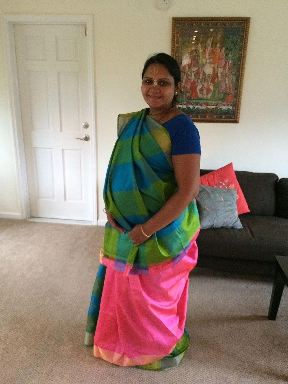 Rina, pregnant, wearing a green and pink saree standing in front of a framed painting of a religious scene.