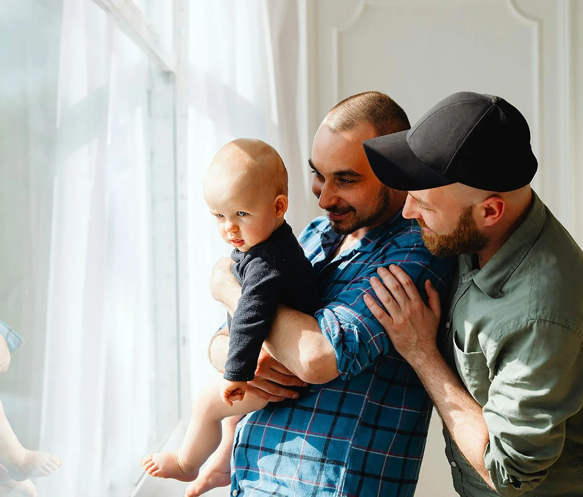 Two parents holding a baby together near a window.