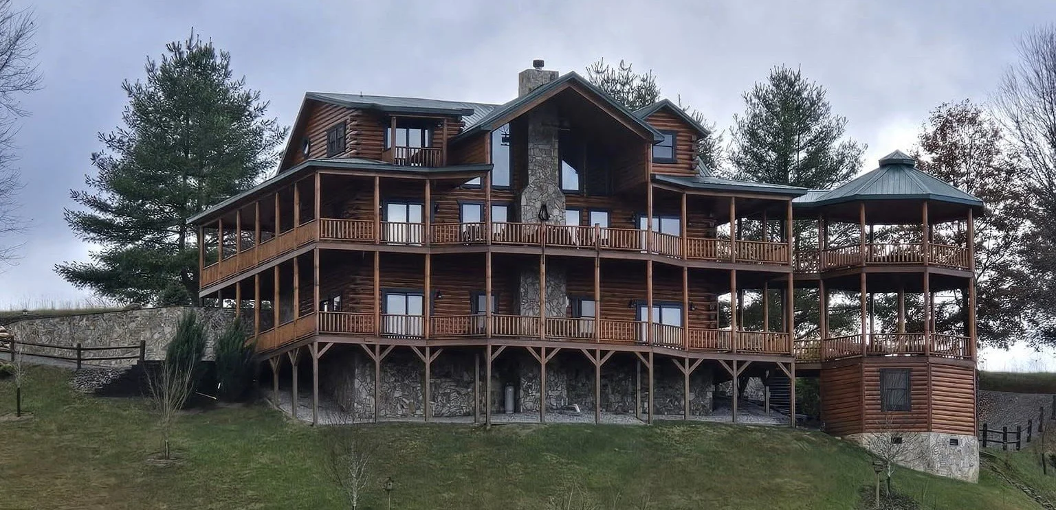 Large wooden house with multiple decks and a stone chimney, situated on a grassy hill with trees in the background.