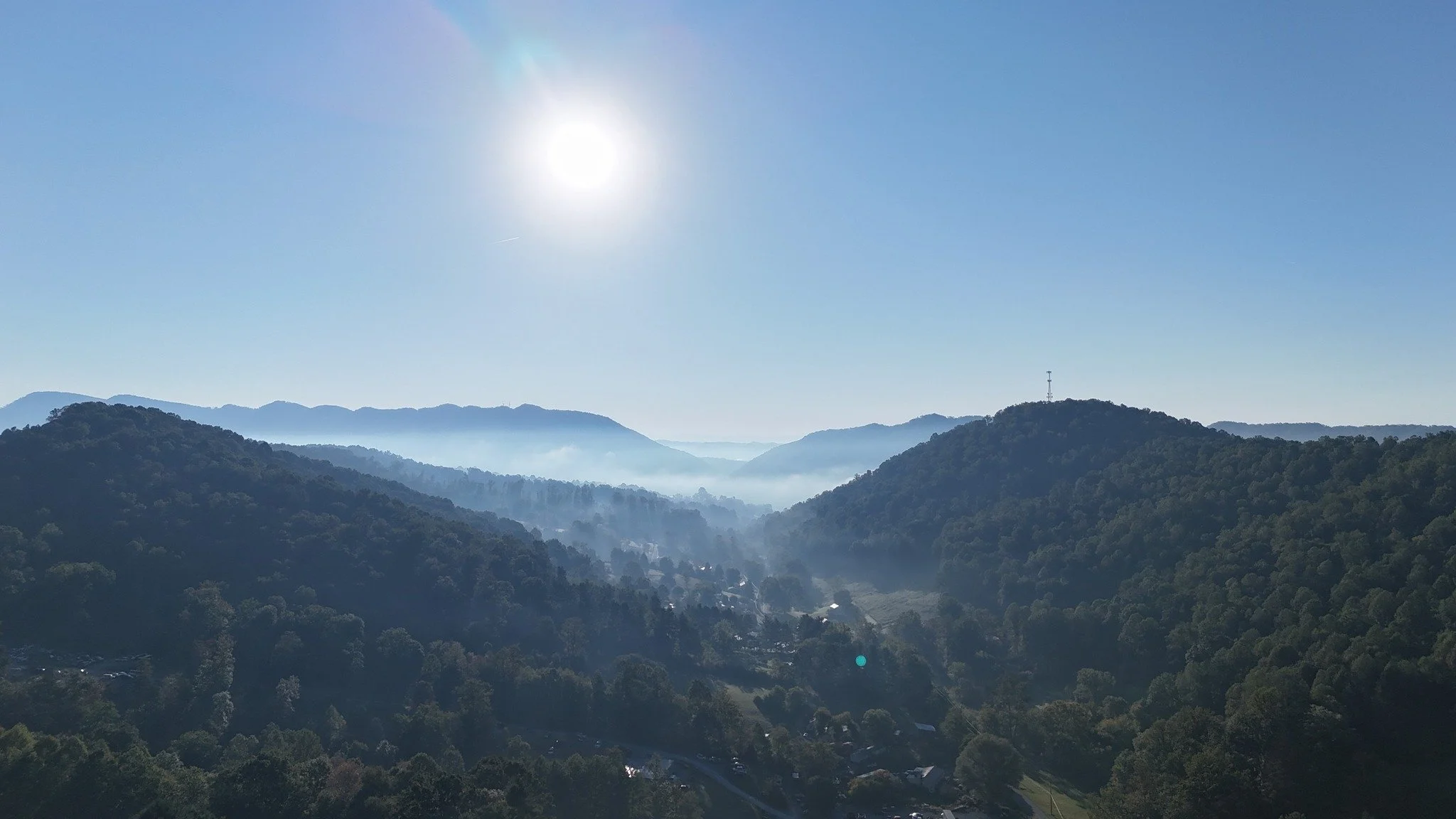 Mountain landscape with forested hills and mist in the valley, bright sun in a clear blue sky.