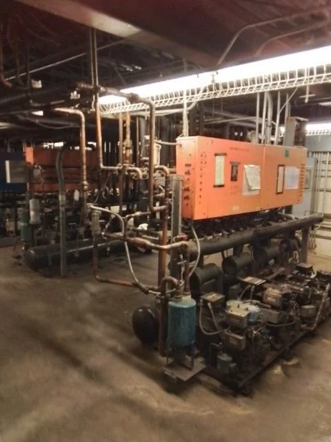 Industrial mechanical room with pipes, valves, and machinery including an orange electrical panel and generators.